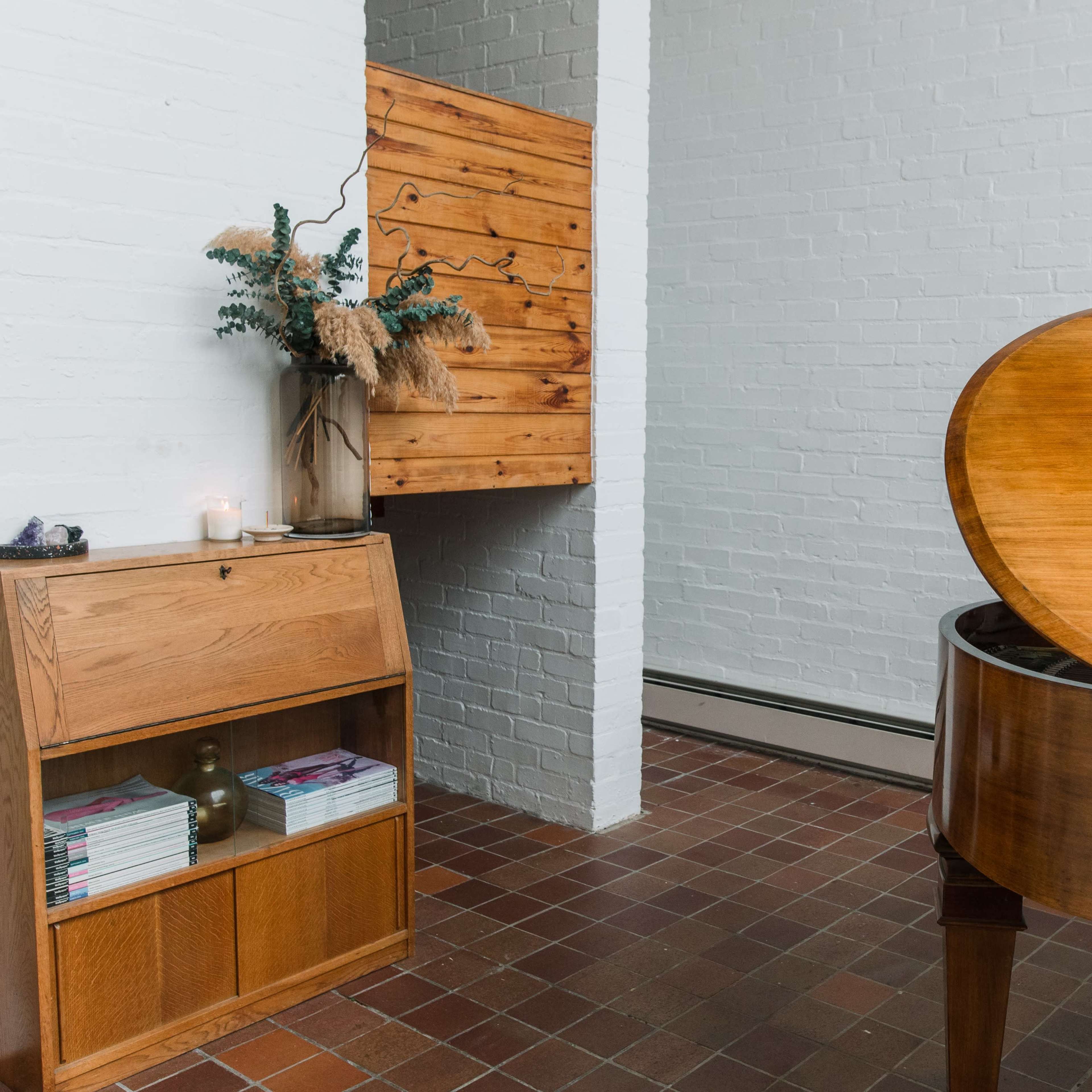 A wooden grand piano beside a minimalist wooden cabinet and a wall-mounted storage unit, set in a bright, sparsely furnished room with white brick walls.