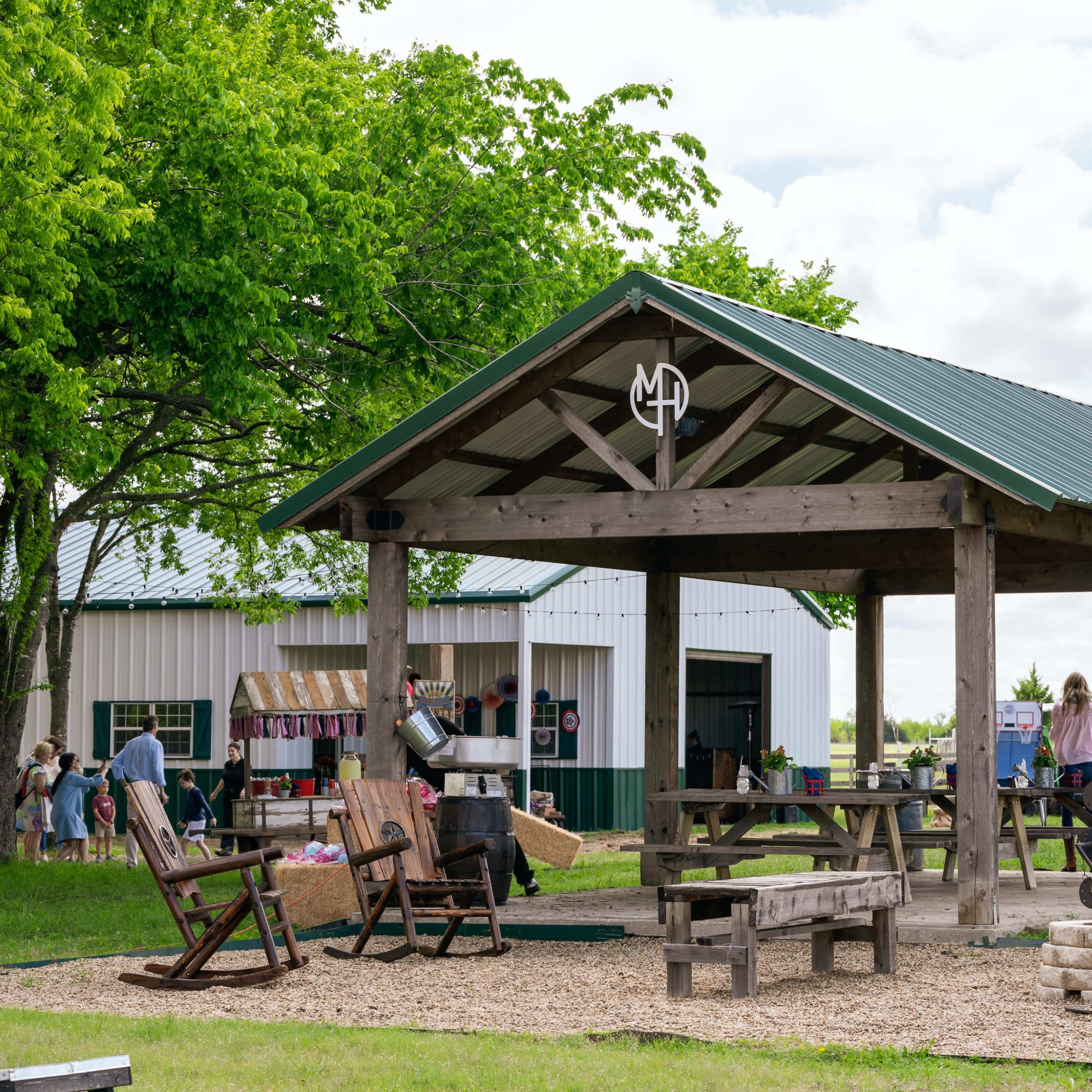 A shaded outdoor pavilion with wooden picnic tables and rocking chairs is set near a barn and grassy area.