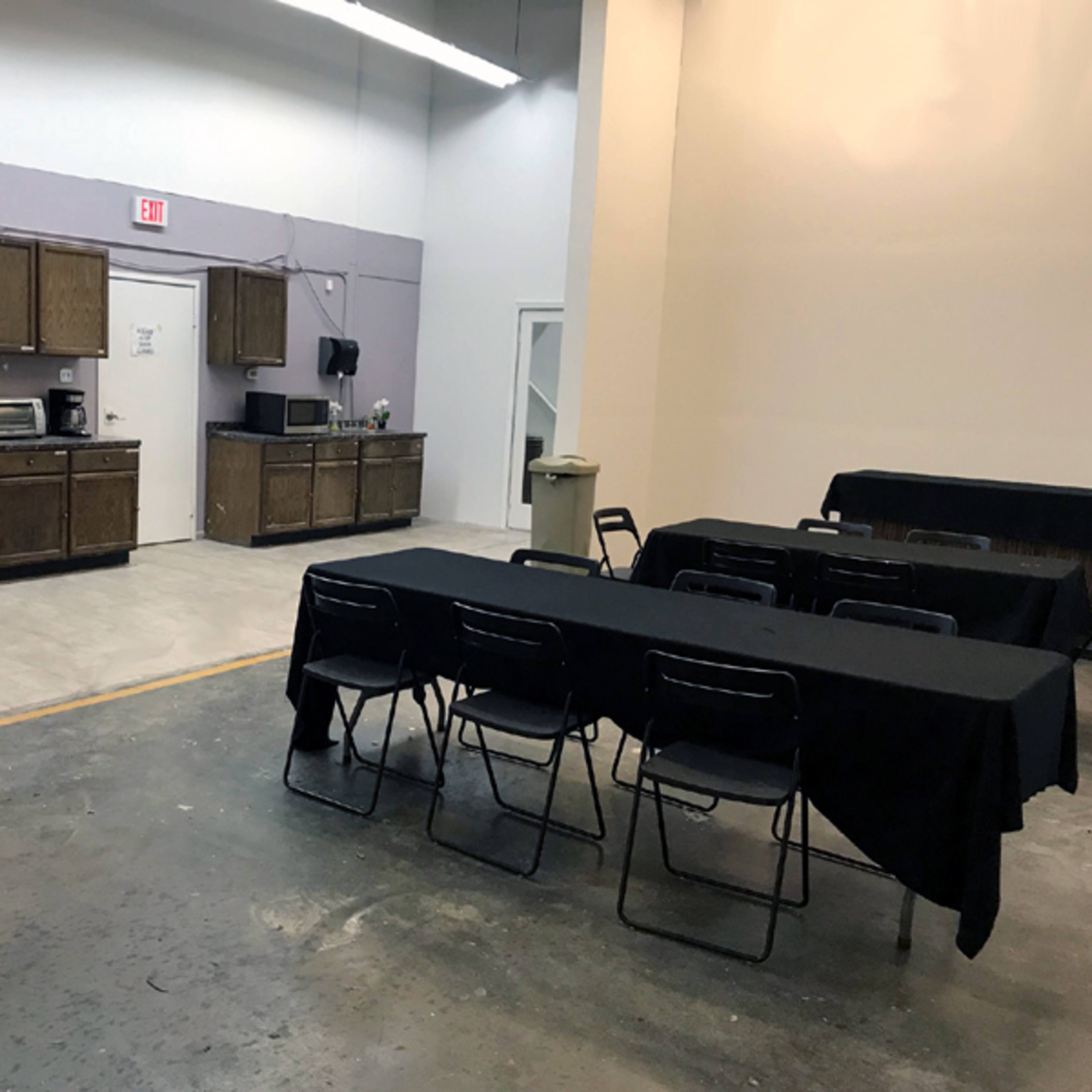 A simple, spacious kitchen area with a refrigerator, cabinets, and several tables covered with black tablecloths.