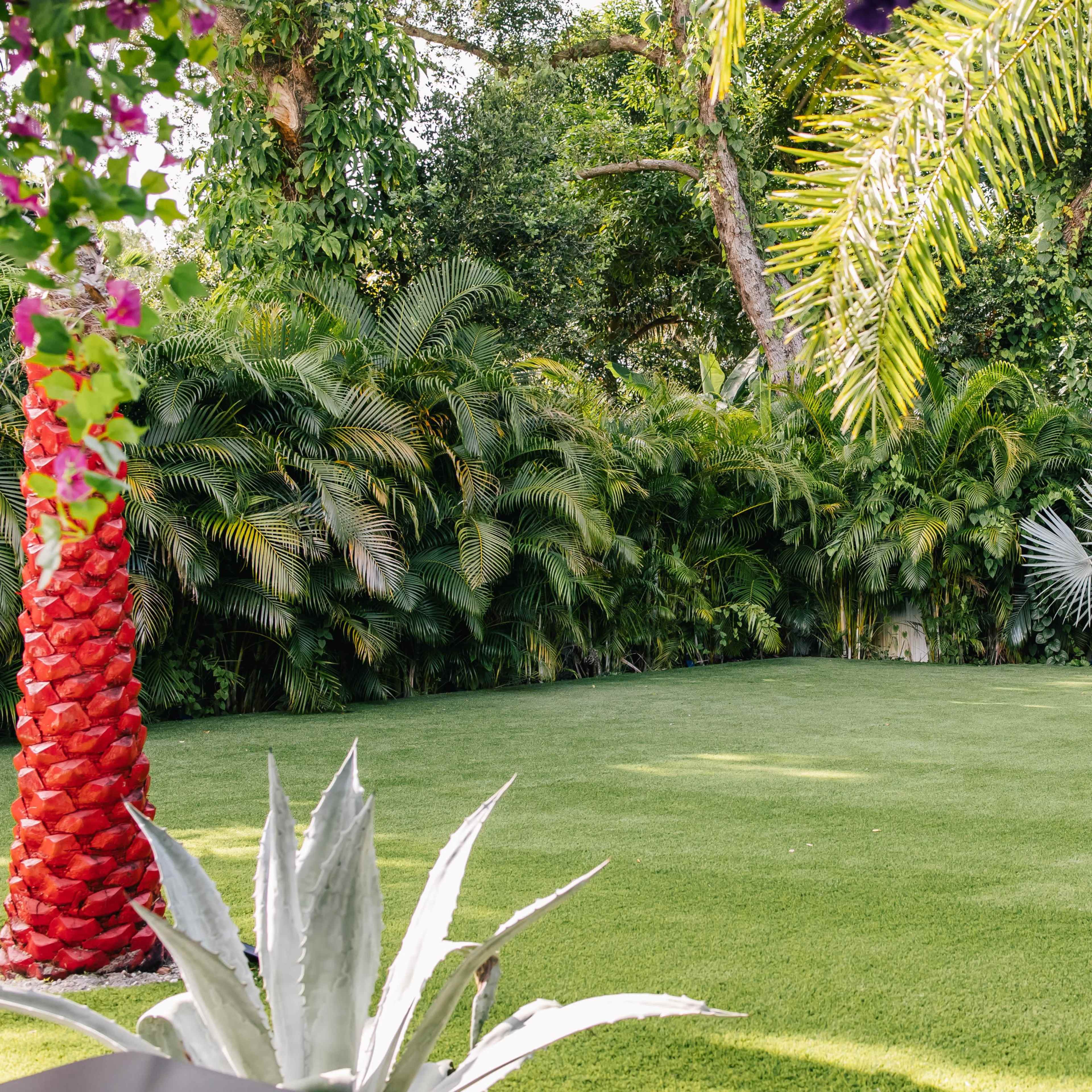 A lush green garden with manicured artificial grass, bordered by tropical plants and palm trees.