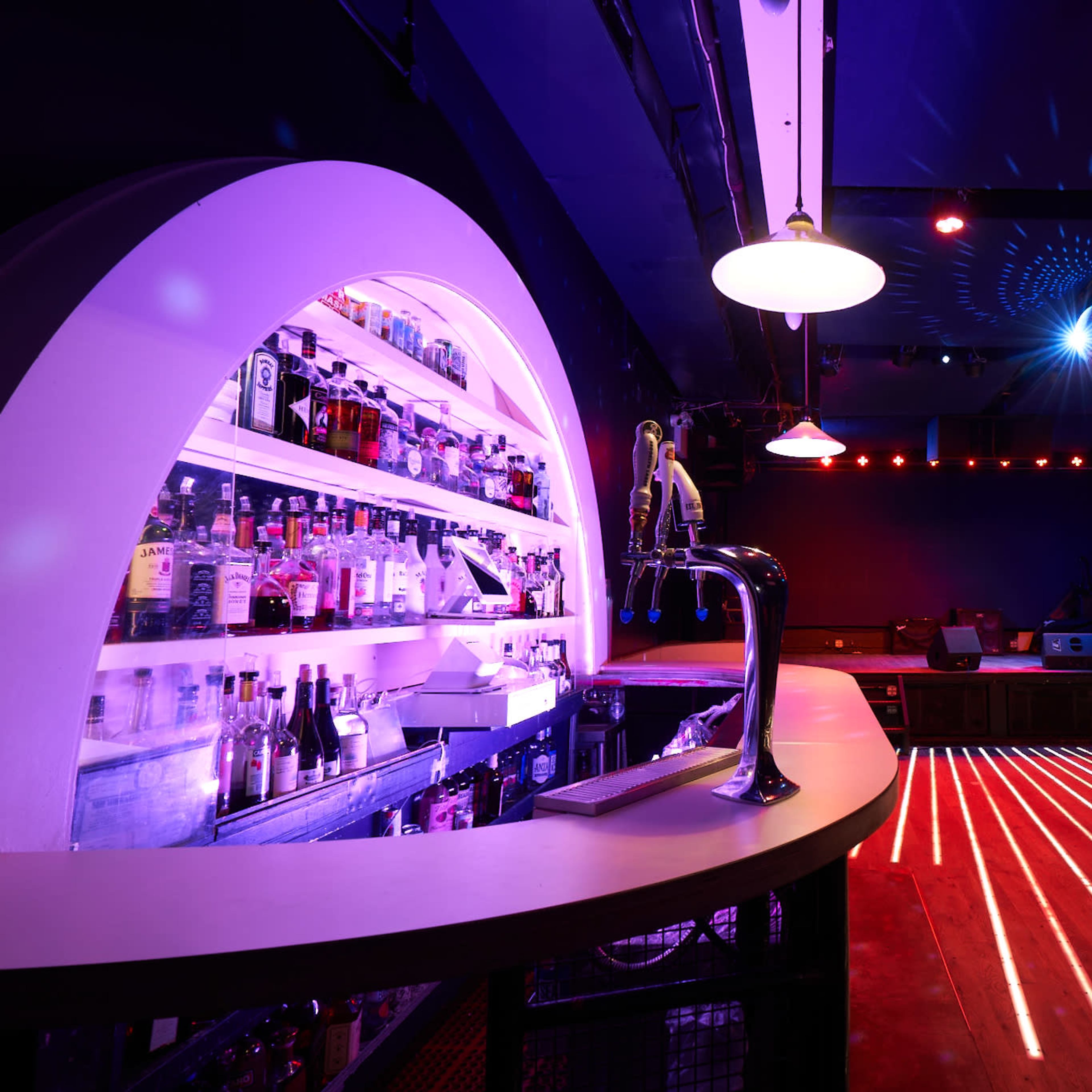 A dimly lit bar with a curved backlit shelf displaying a variety of bottles and a counter with an illuminated surface, accented by colorful lighting effects on the walls and floor.