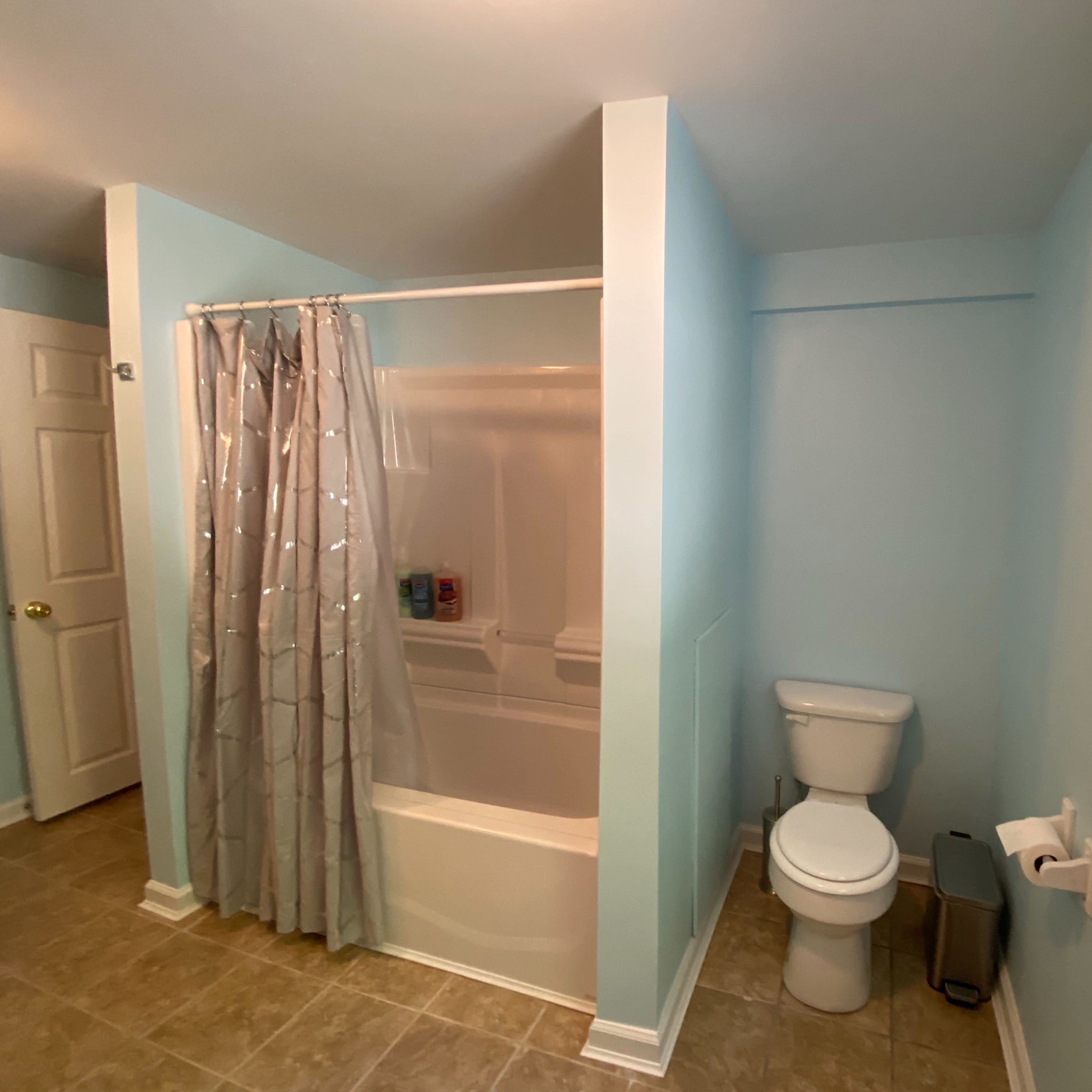 A bathroom features a light blue color scheme, a shower curtain separating a bathtub from the rest of the space, a toilet, and a small waste bin.