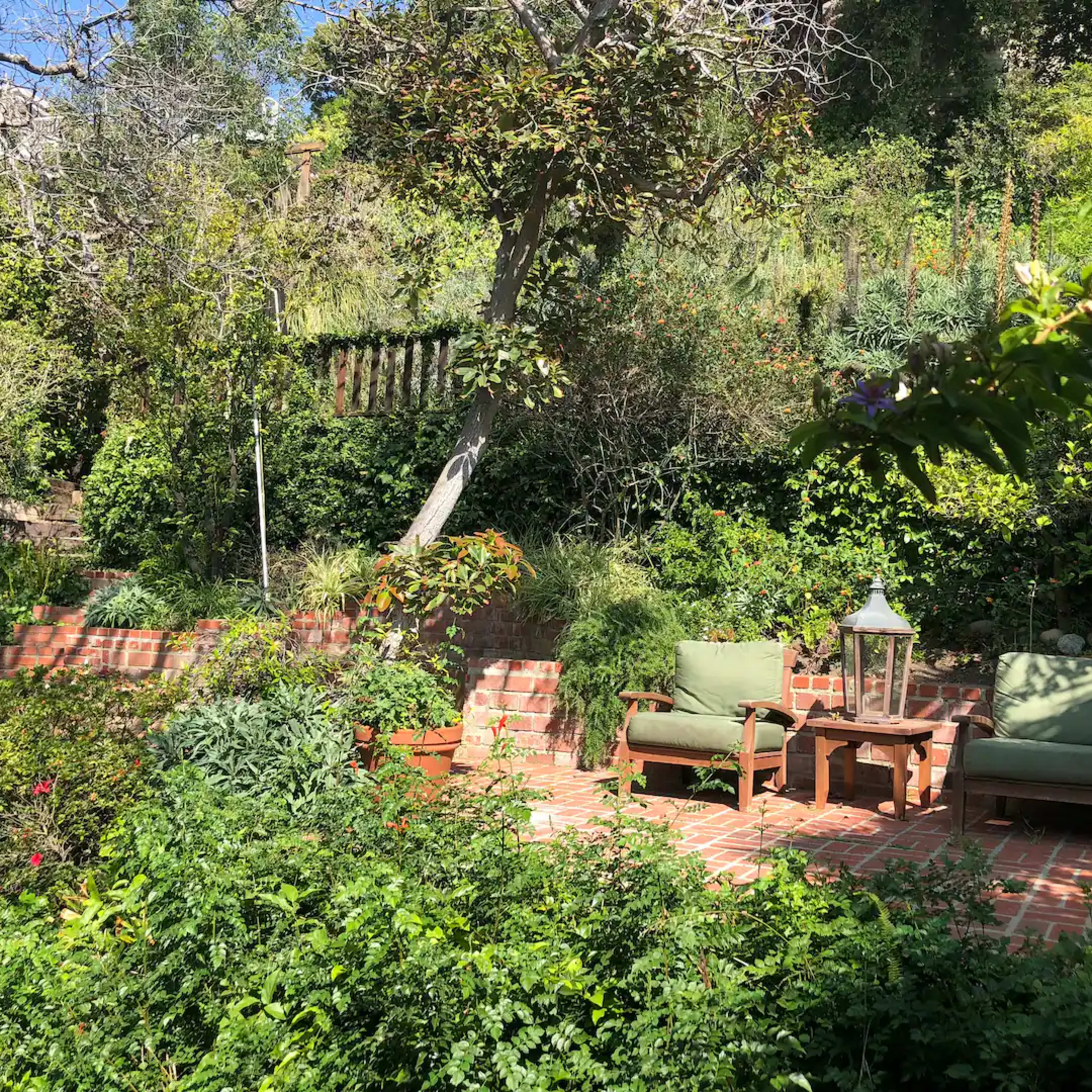 A brick patio area with two green chairs and a small table, surrounded by lush greenery and trees.