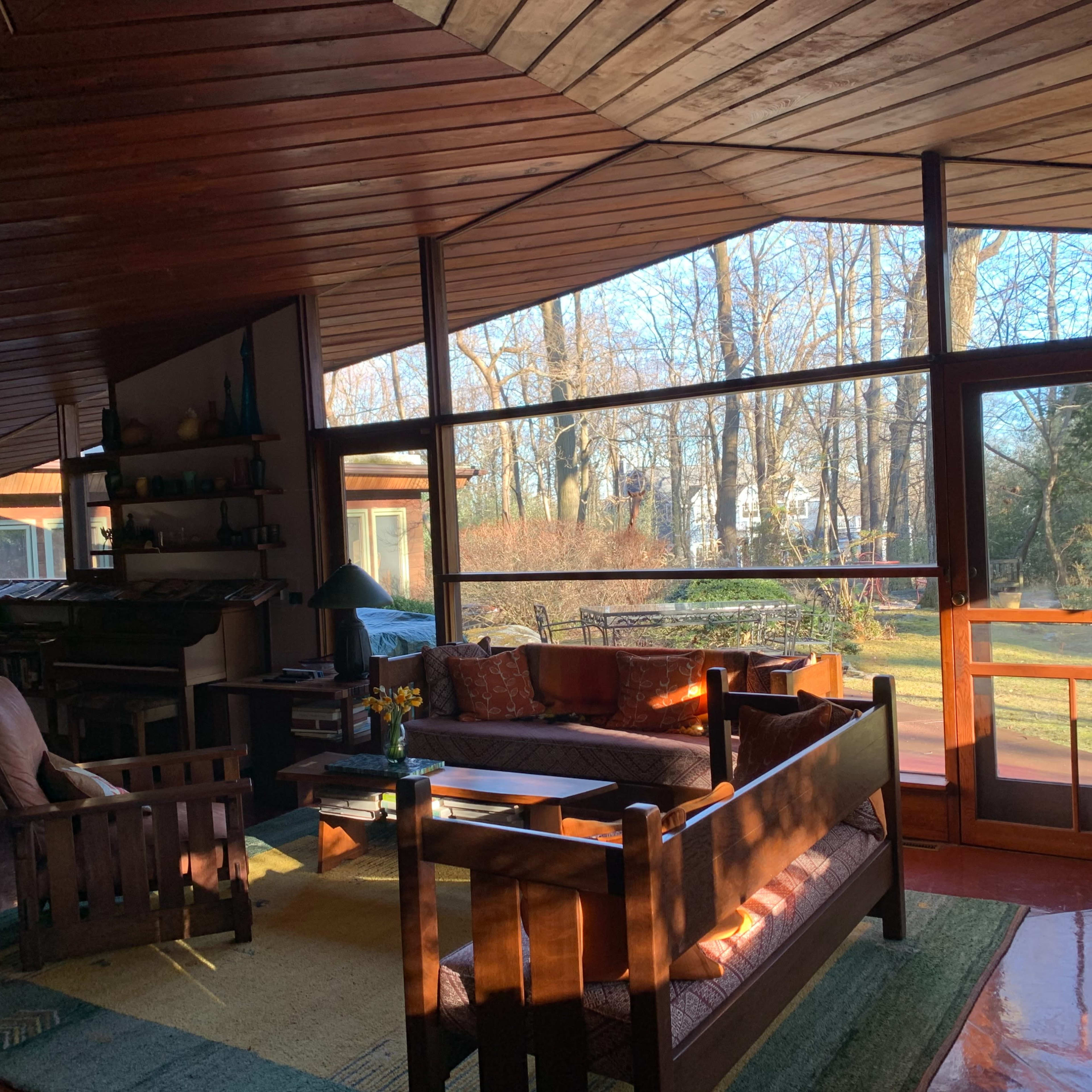 A spacious living room with wooden walls and ceiling, filled with natural light and featuring a large glass window that opens to a view of trees outside.