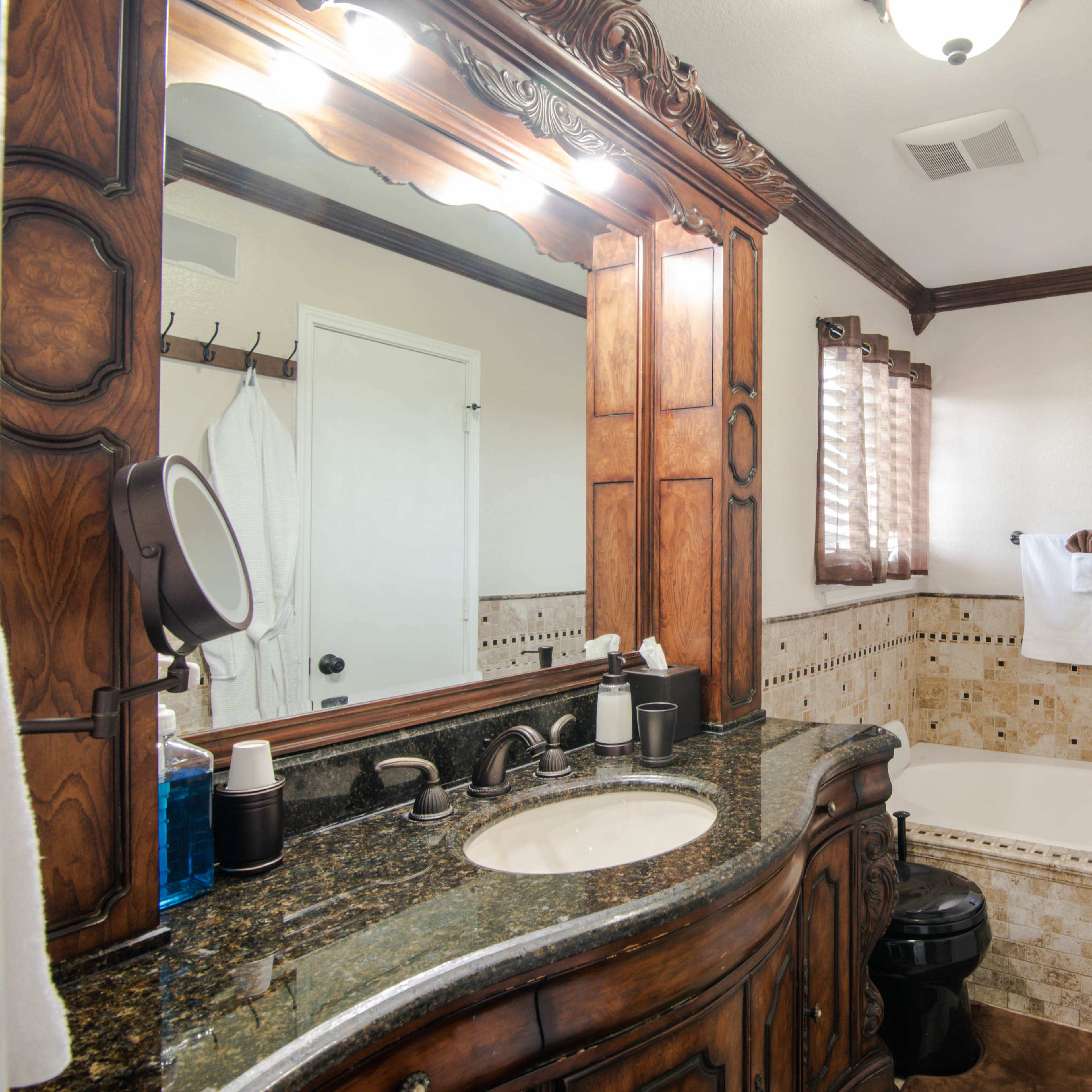 A bathroom features a dark wooden vanity with a marble countertop, a large mirror with bright lights, and a freestanding bathtub surrounded by tiled walls.