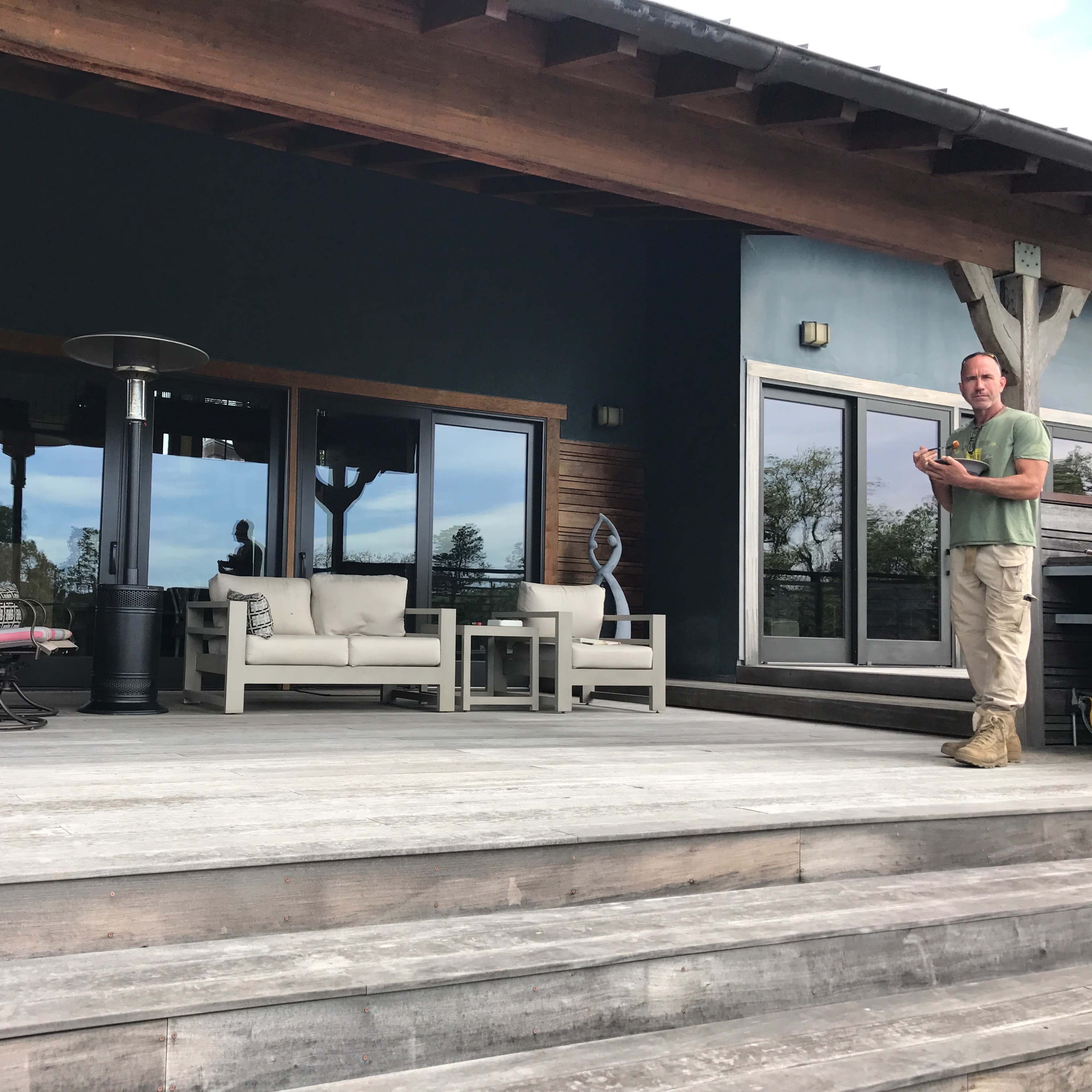 A man stands on the wooden steps of a modern house with large glass doors, accompanied by outdoor furniture and a patio heater.