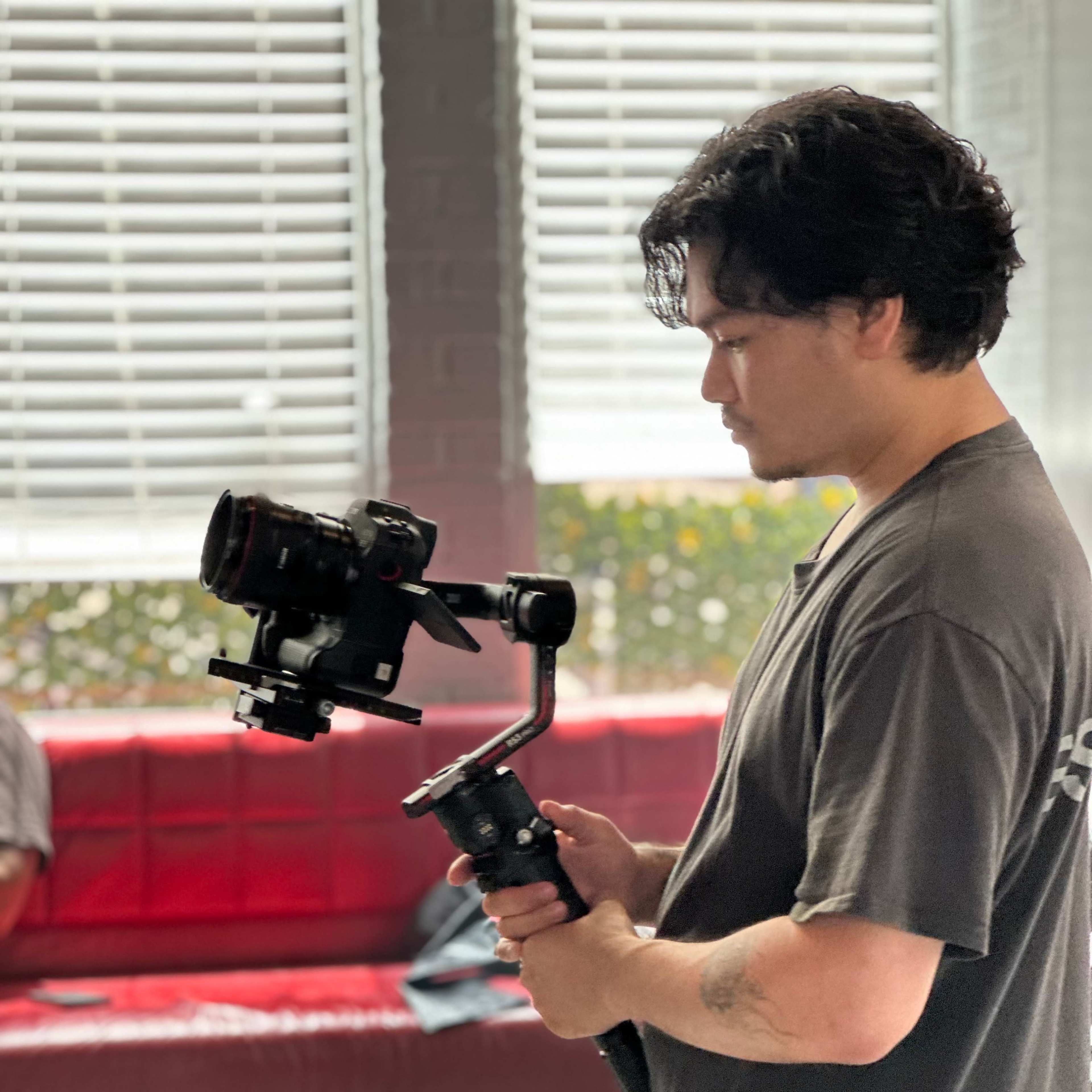 A man stands holding a camera stabilizer in a brightly lit room with large windows and a red couch in the background.