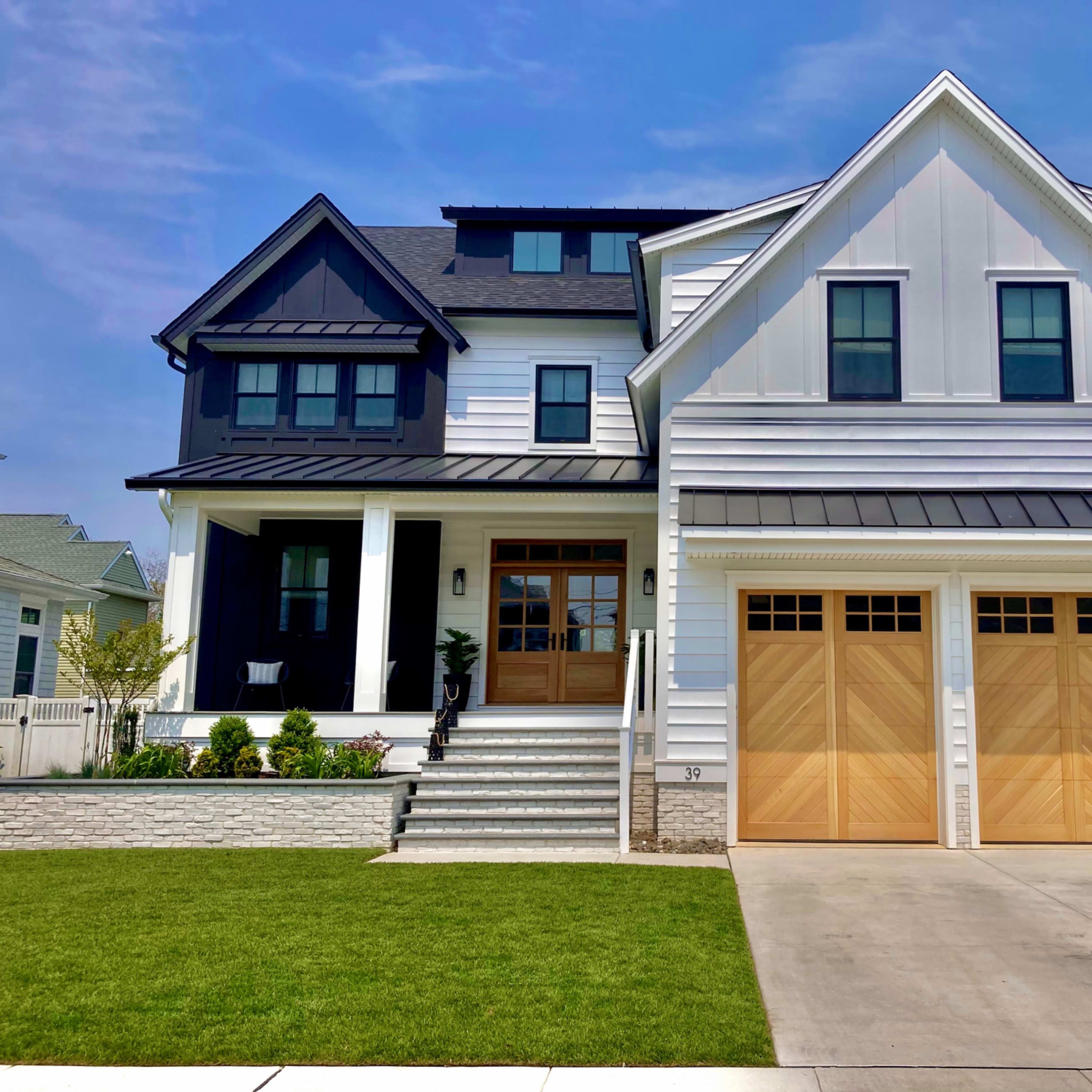 A large, modern two-story house with a mix of black and white siding, a front porch with columns, and a driveway leading to a garage with wooden doors.