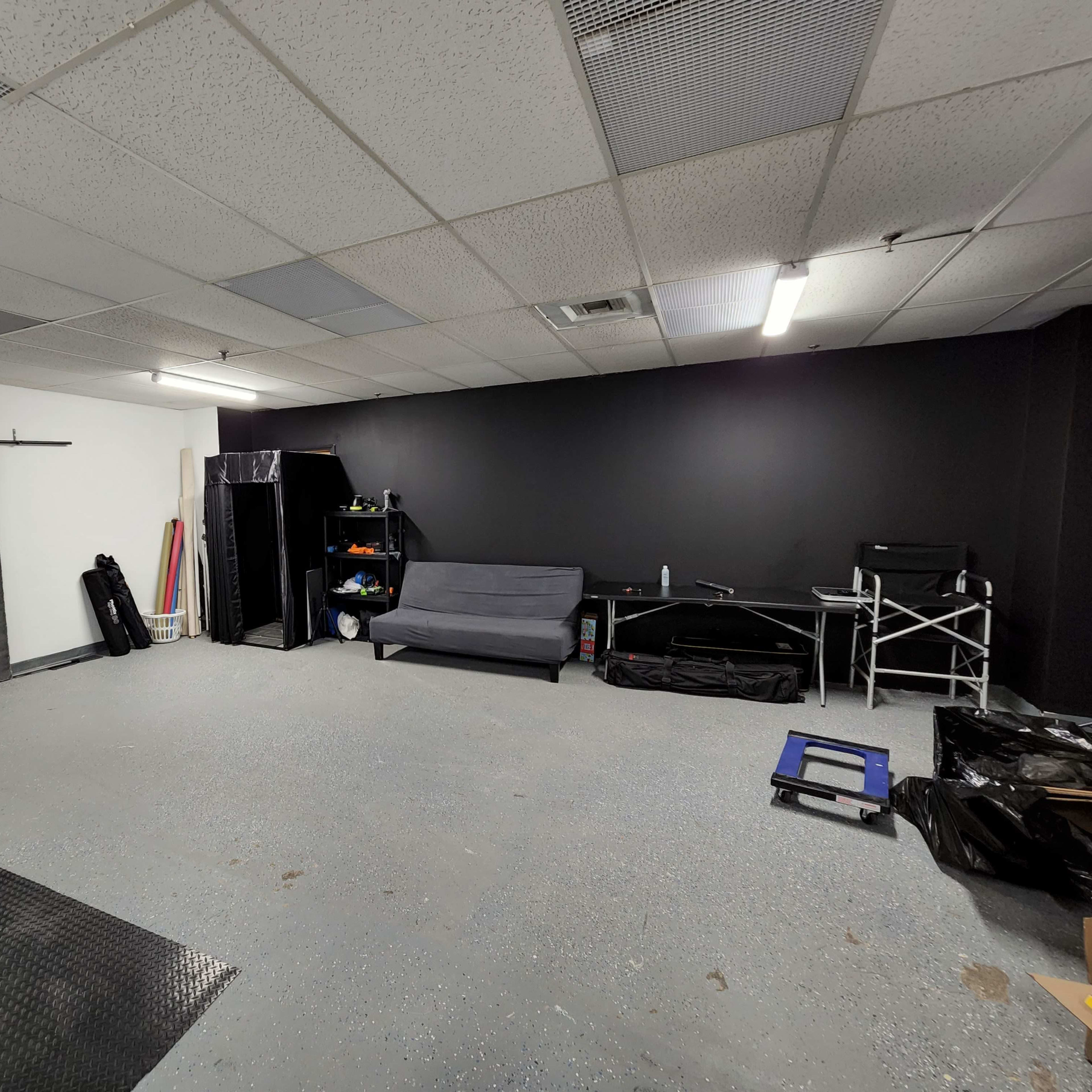 A spacious, empty room with a gray concrete floor, a black wall, and minimal furniture, including a gray couch and a few storage items.