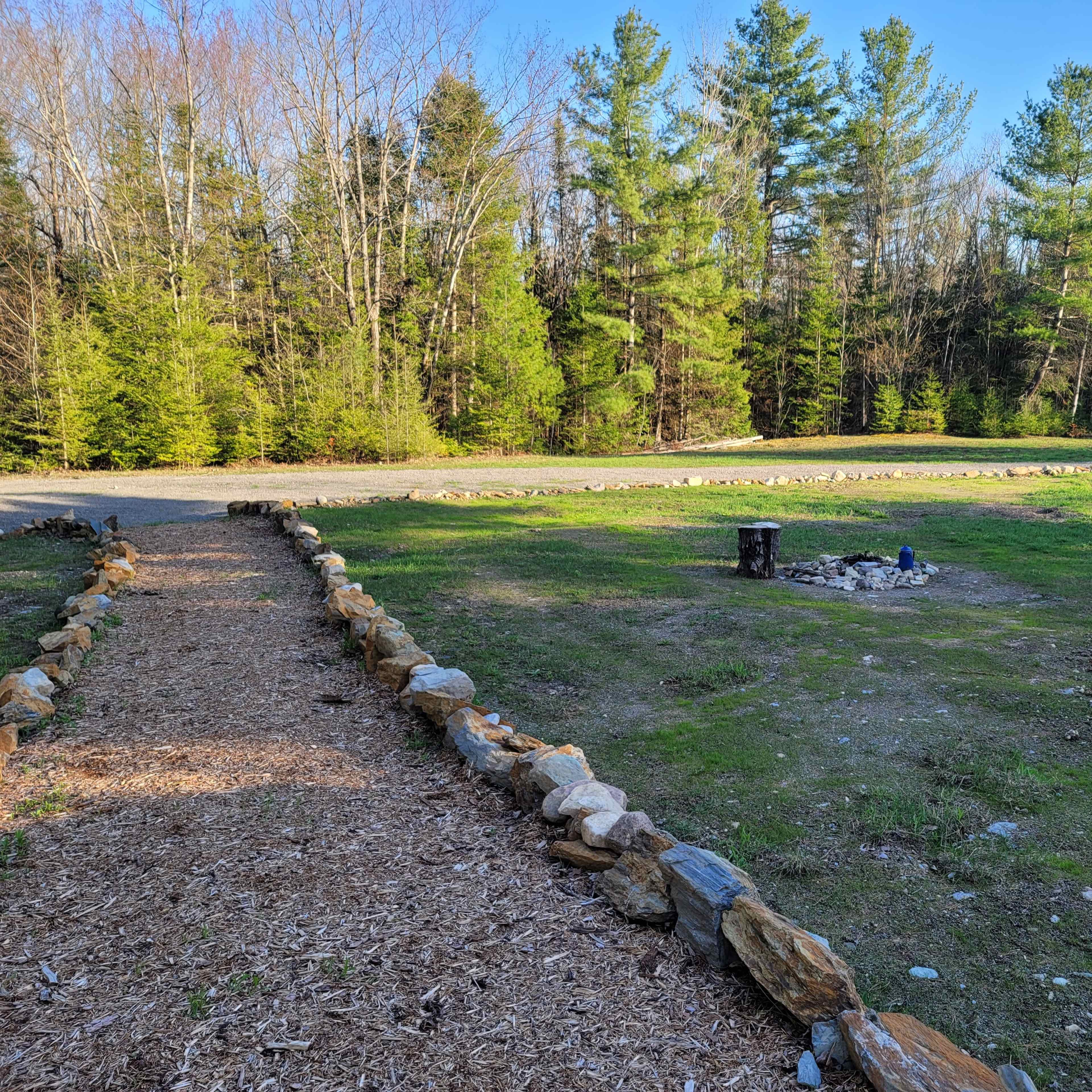 A gravel pathway lined with stones leads to a fire pit surrounded by grassy land and trees in the background.