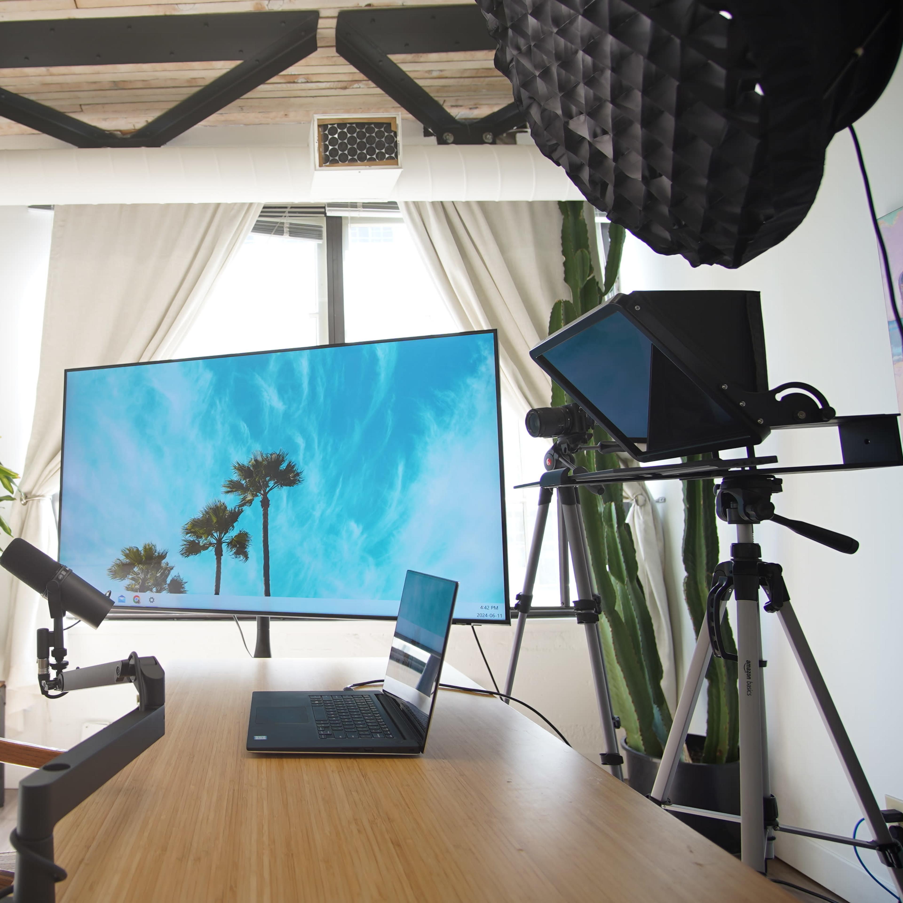 A workspace features a large monitor displaying palm trees, a laptop on a wooden desk, and a microphone alongside a camera setup.