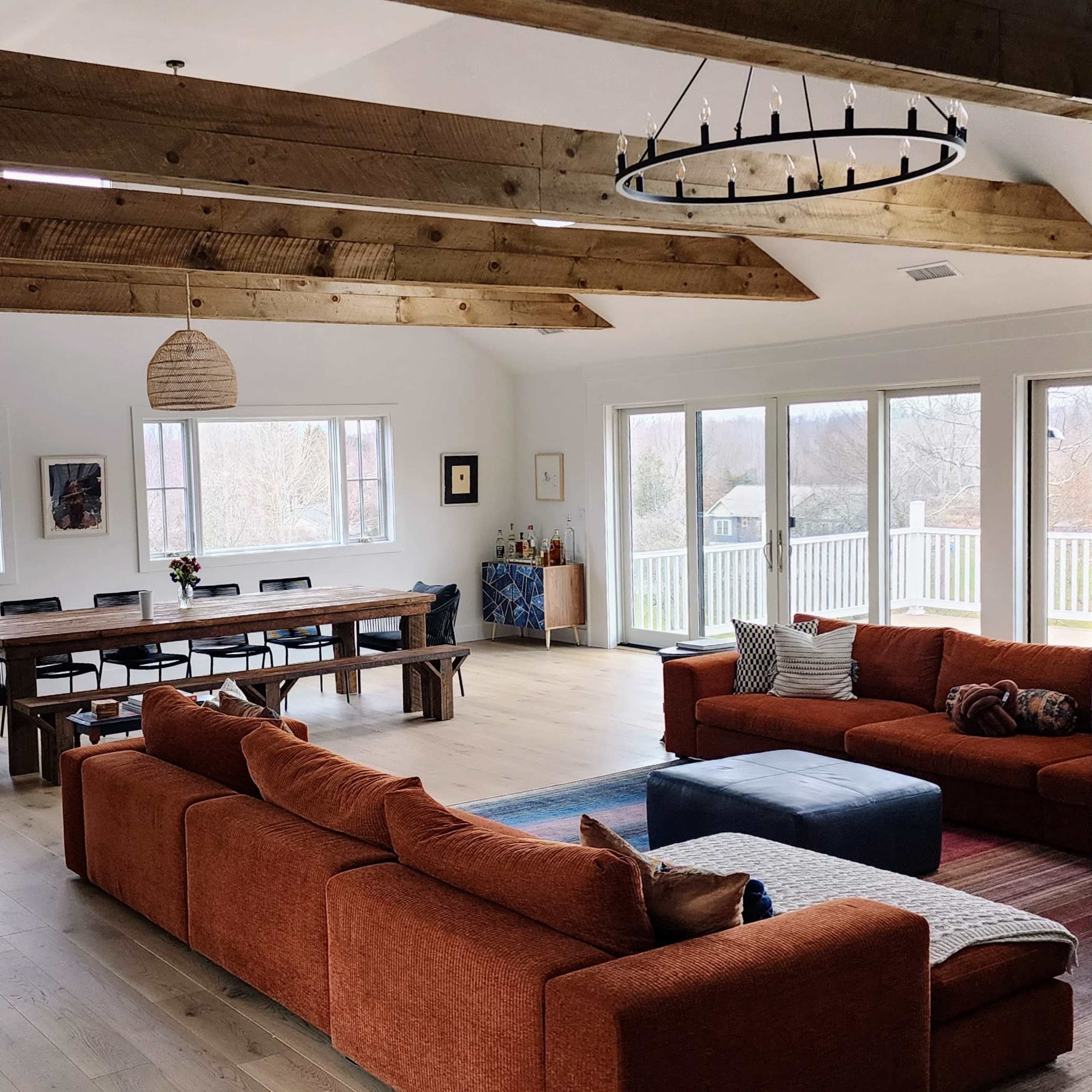 A spacious living area with wooden beams, a large table, multiple seating options, and large windows overlooking a green landscape.