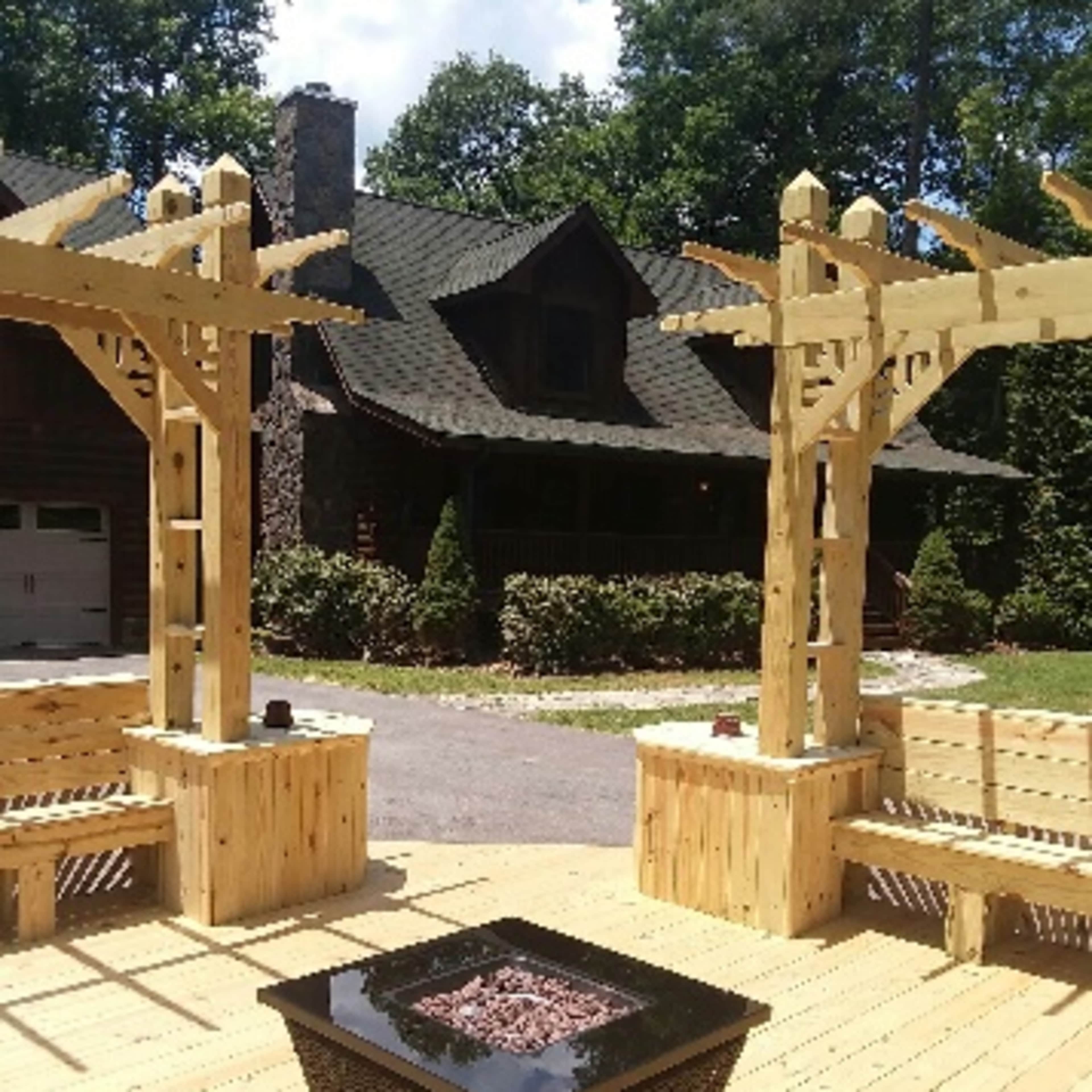 A wooden deck with a fire pit surrounded by benches and two wooden pergolas, set against a house and trees in the background.