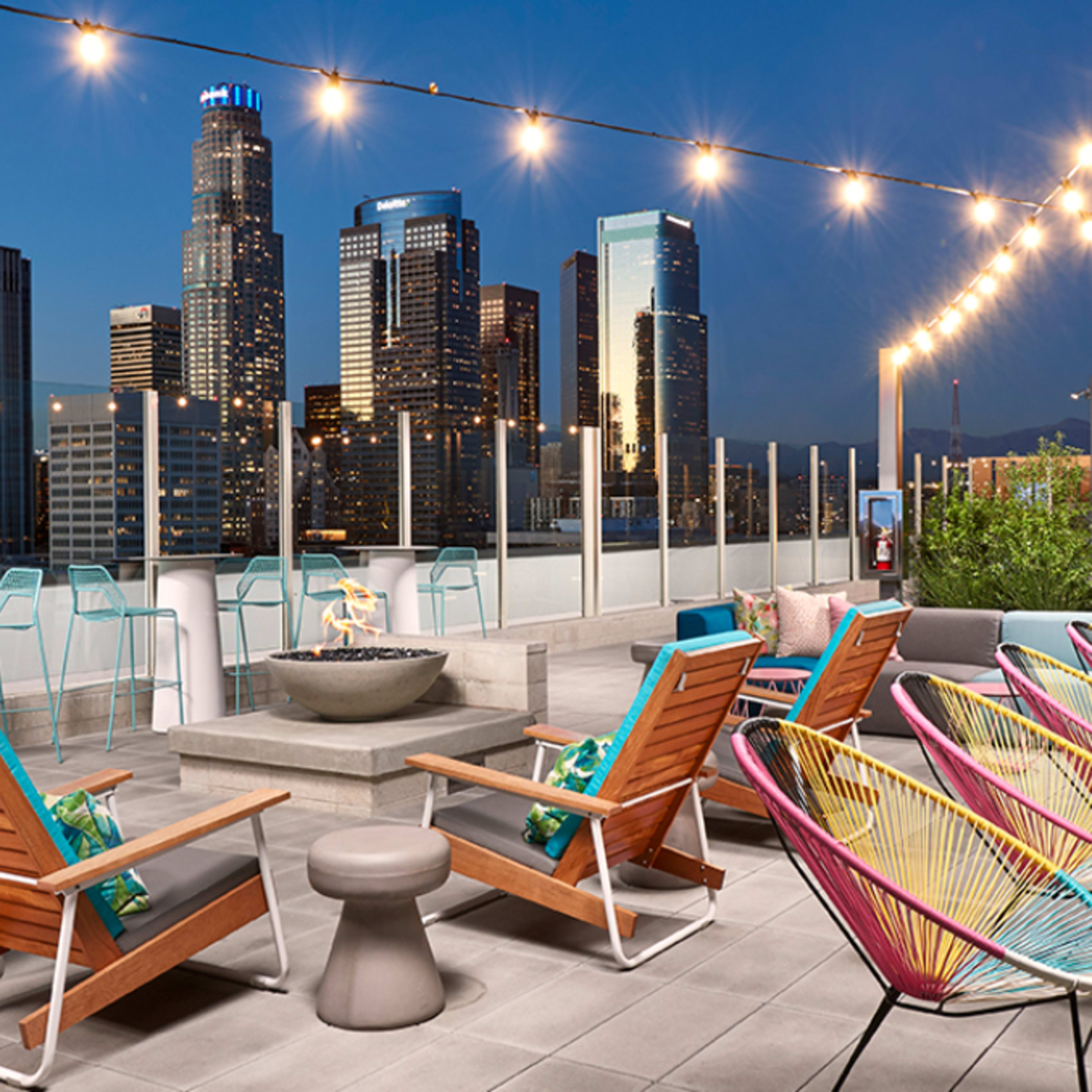 A rooftop terrace with colorful seating, a fire pit, and a skyline view of tall buildings illuminated at dusk.