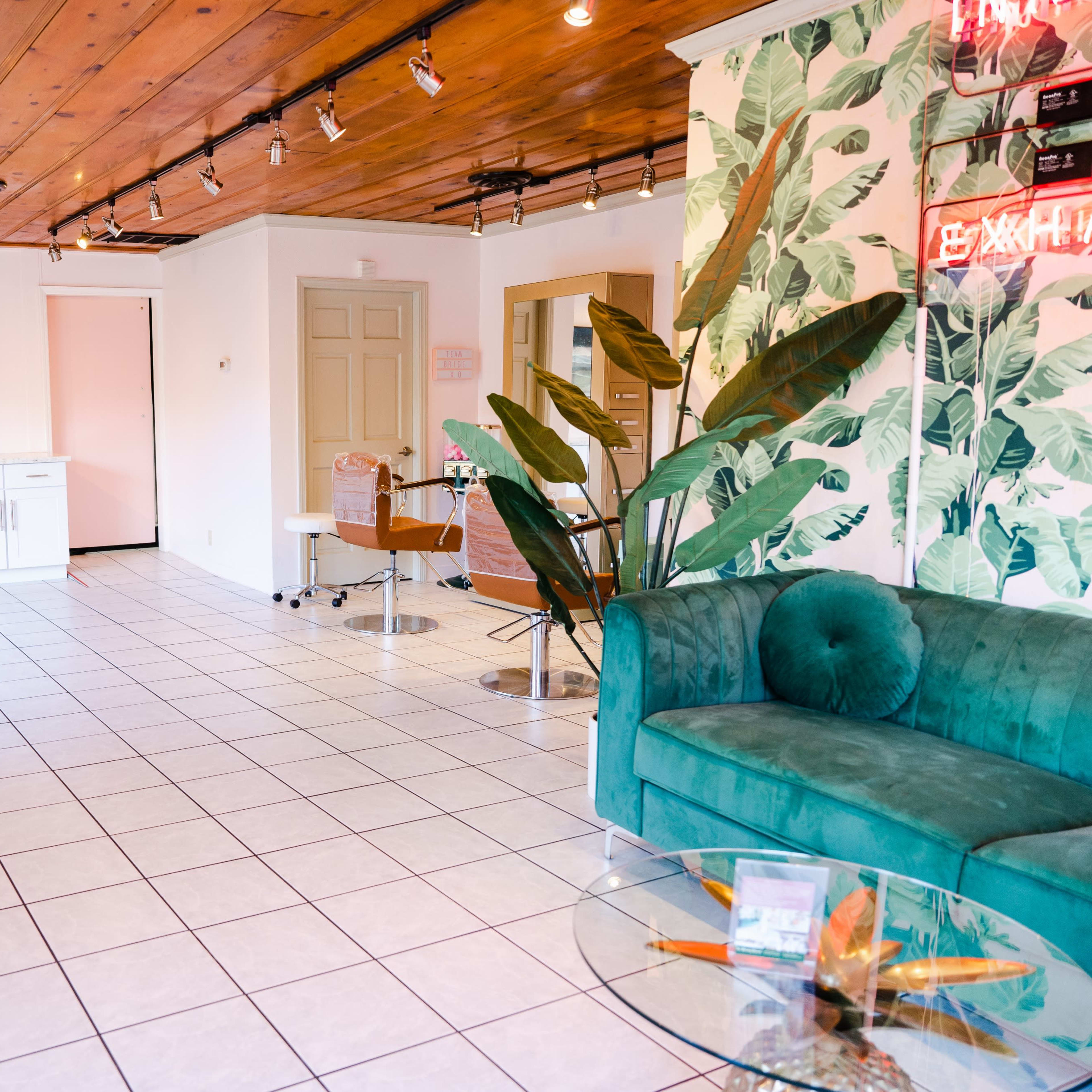 A salon interior with tiled flooring, a green sofa, a coffee table, and a kitchenette area against a backdrop of tropical-themed wallpaper.