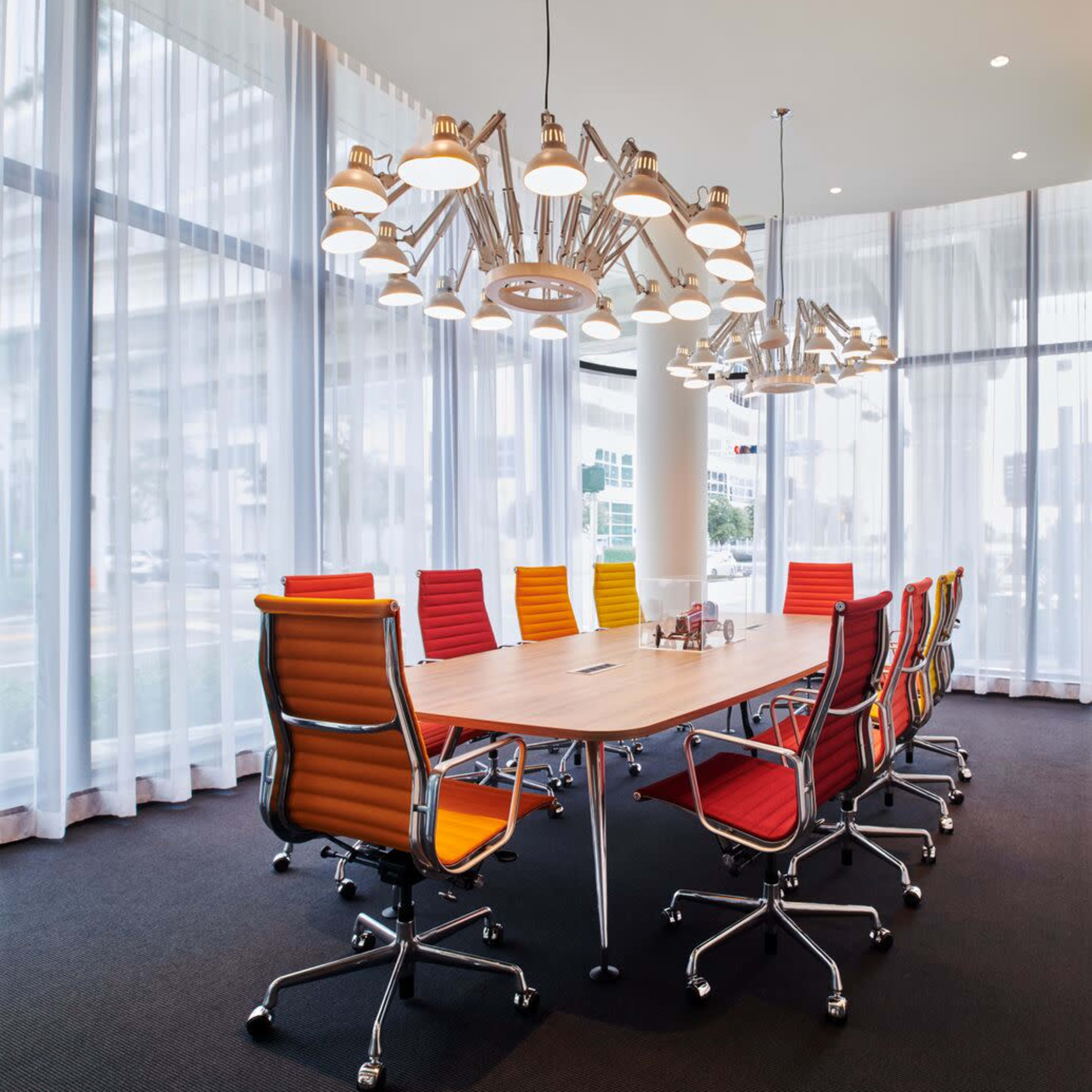 A modern conference room features a large wooden table surrounded by colorful chairs and an overhead light fixture.
