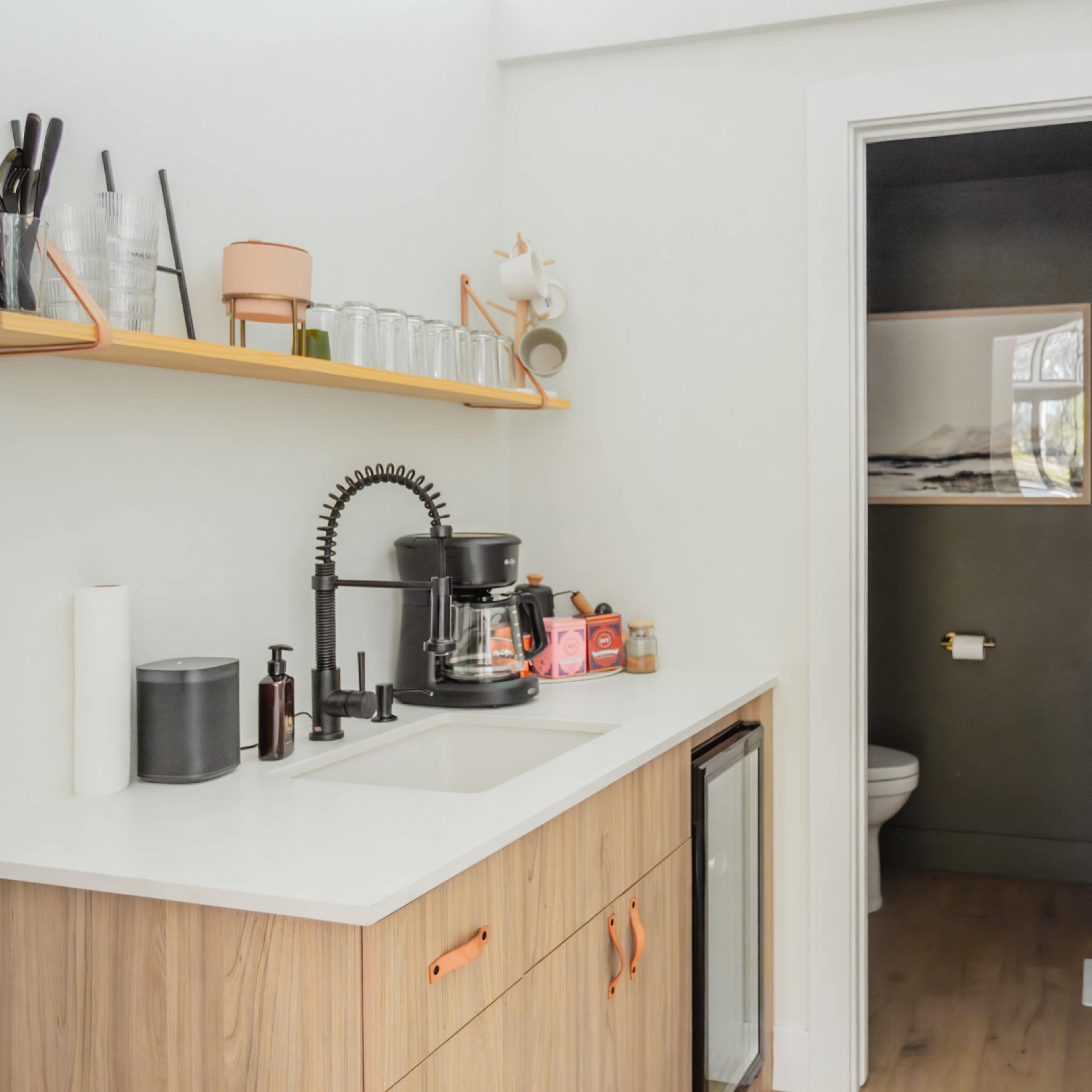 A modern kitchenette with a sink, coffee maker, and open shelving displaying kitchenware, adjacent to a small restroom.