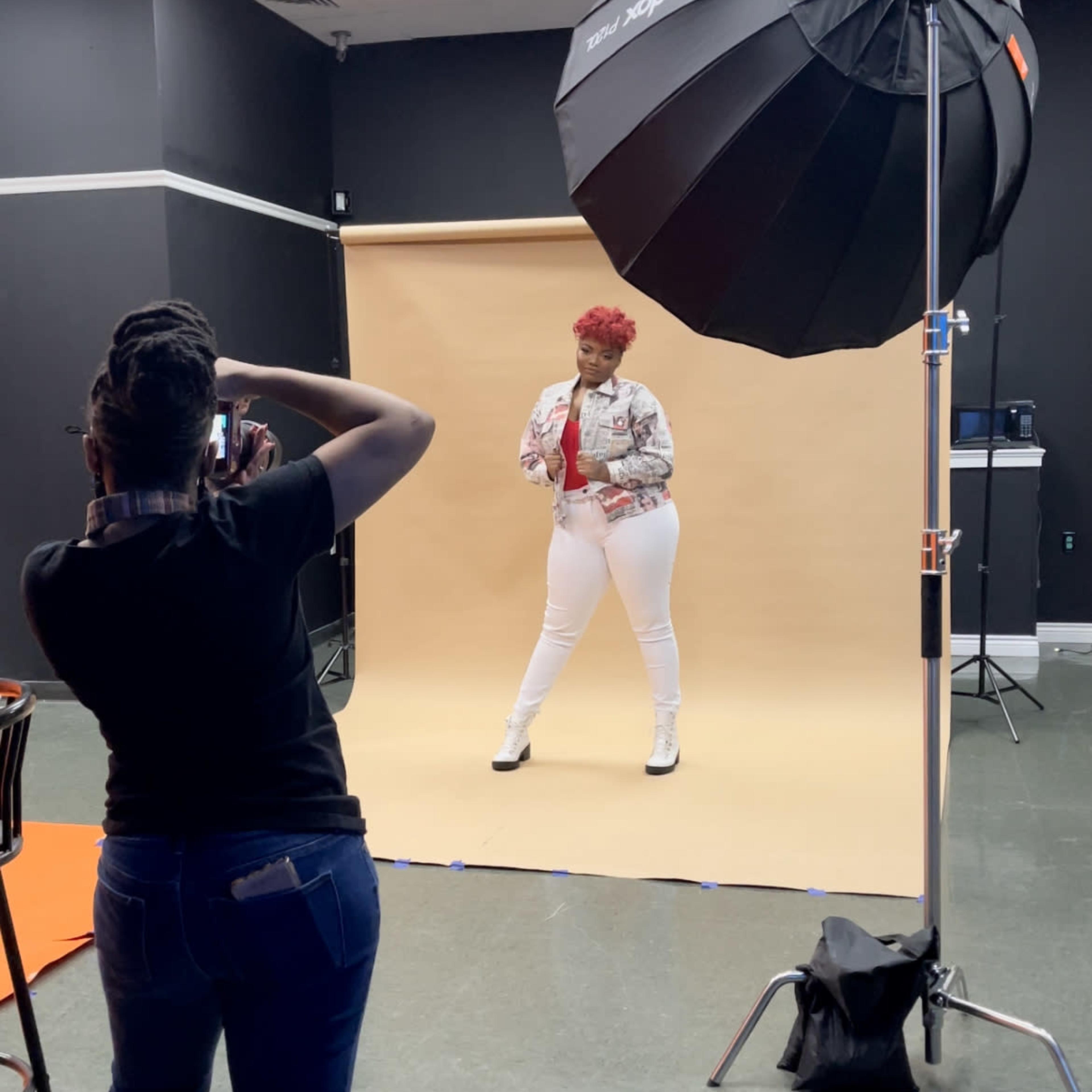 A model poses in front of a beige backdrop while a photographer captures the shot under a studio light.