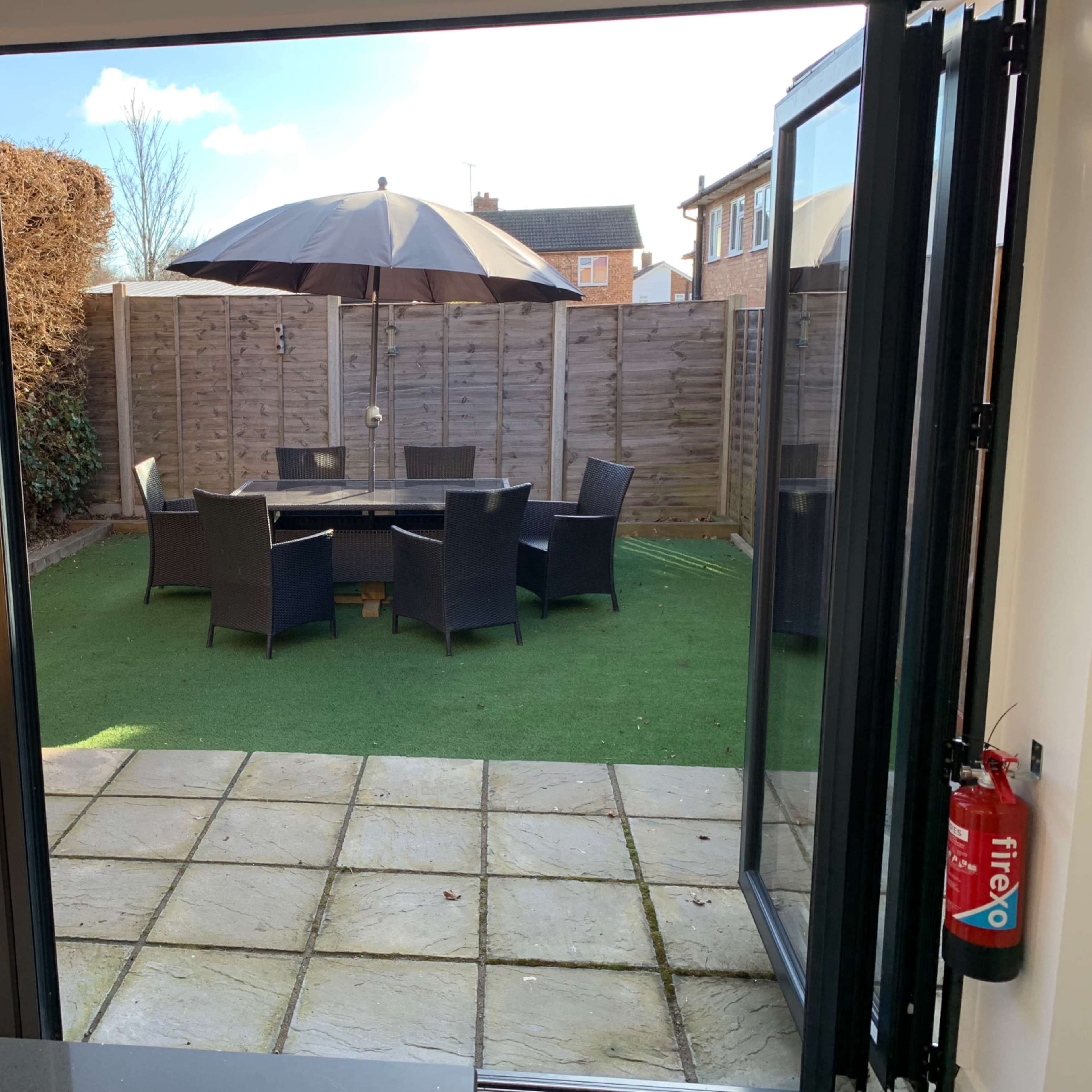 A patio area features a table and chairs set under an umbrella, surrounded by a fenced yard with artificial grass.