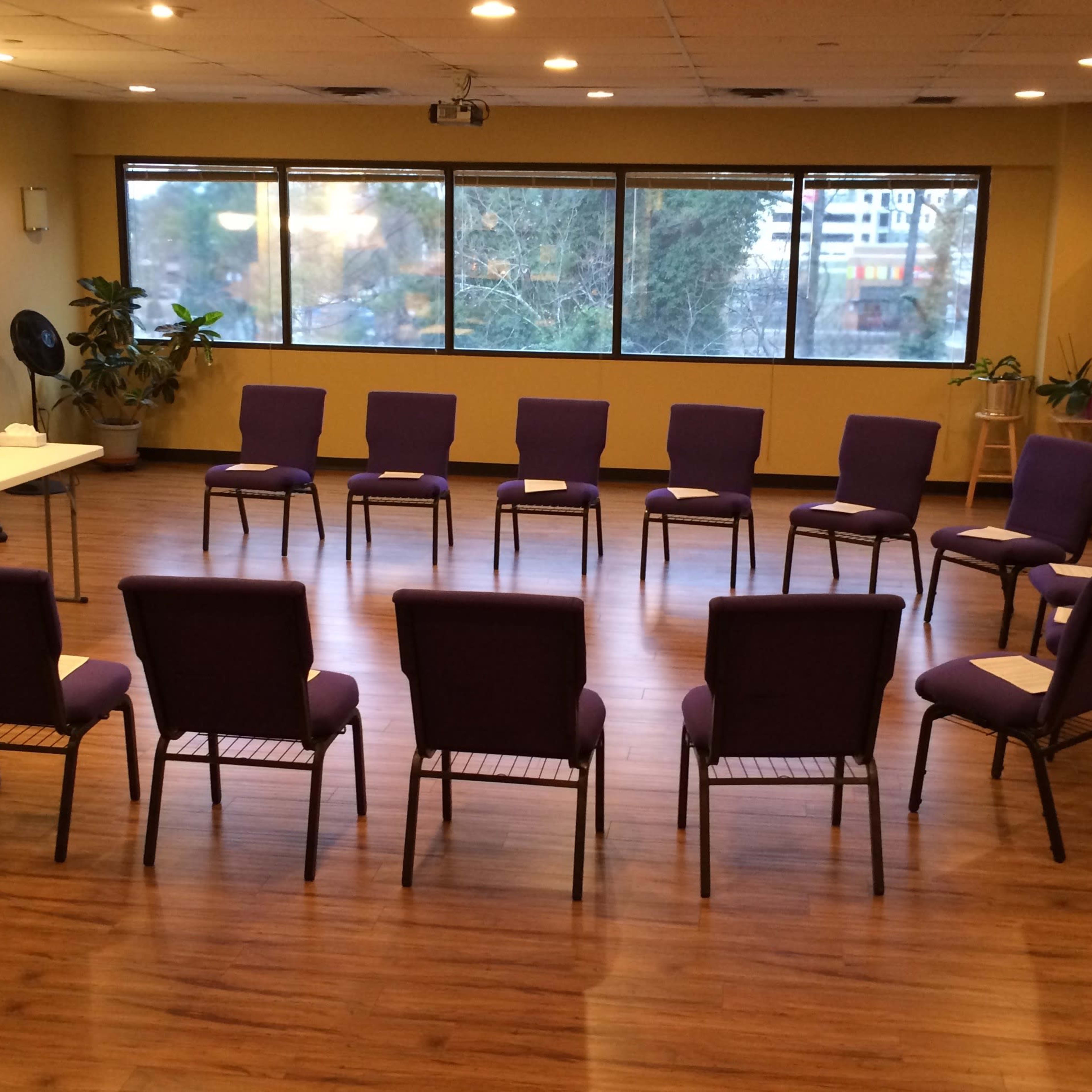 A circular arrangement of purple chairs is set up in a spacious room with large windows and wood flooring.