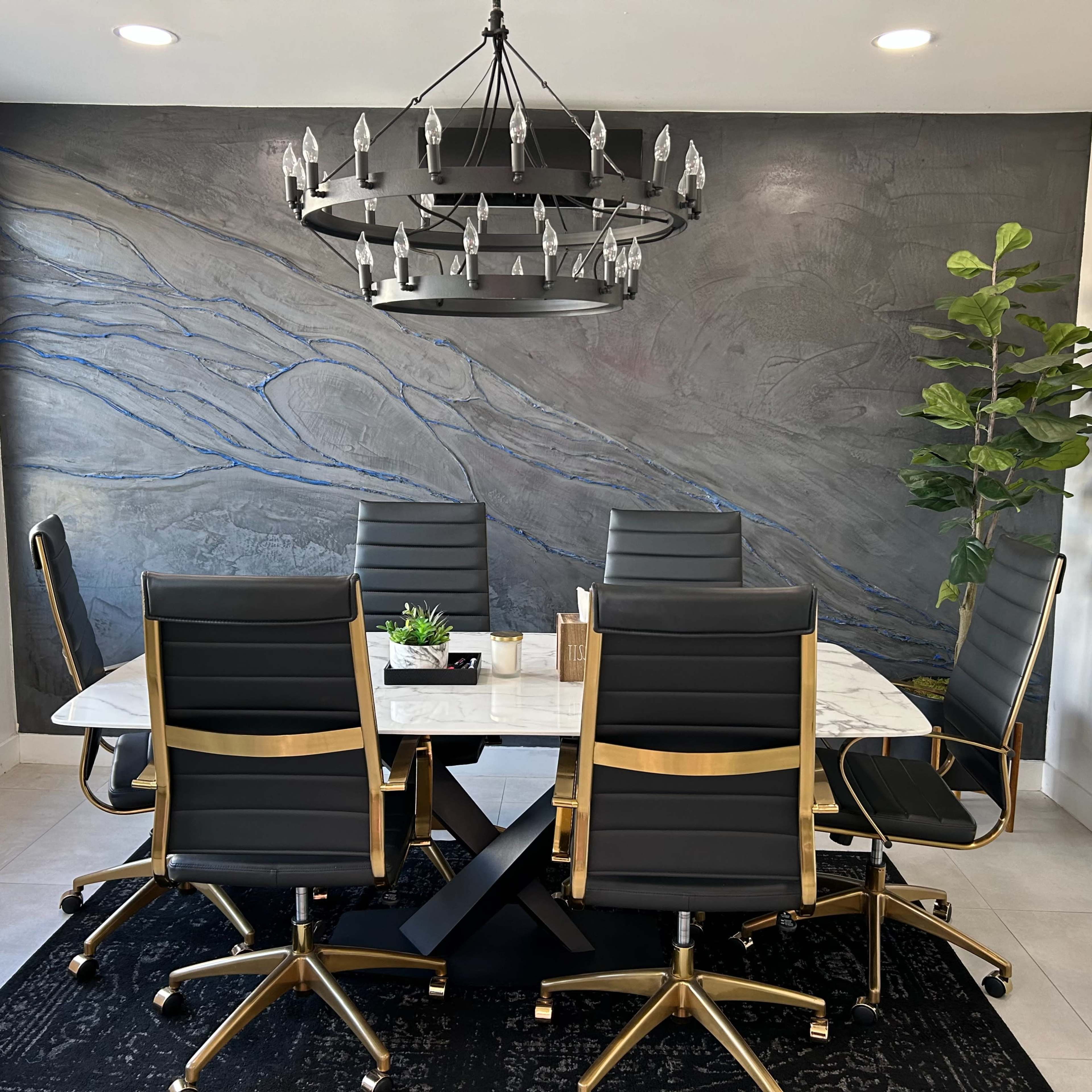 A modern conference room features a large marble table surrounded by sleek black chairs, with a chandelier above and a textured gray wall adorned with blue accents.