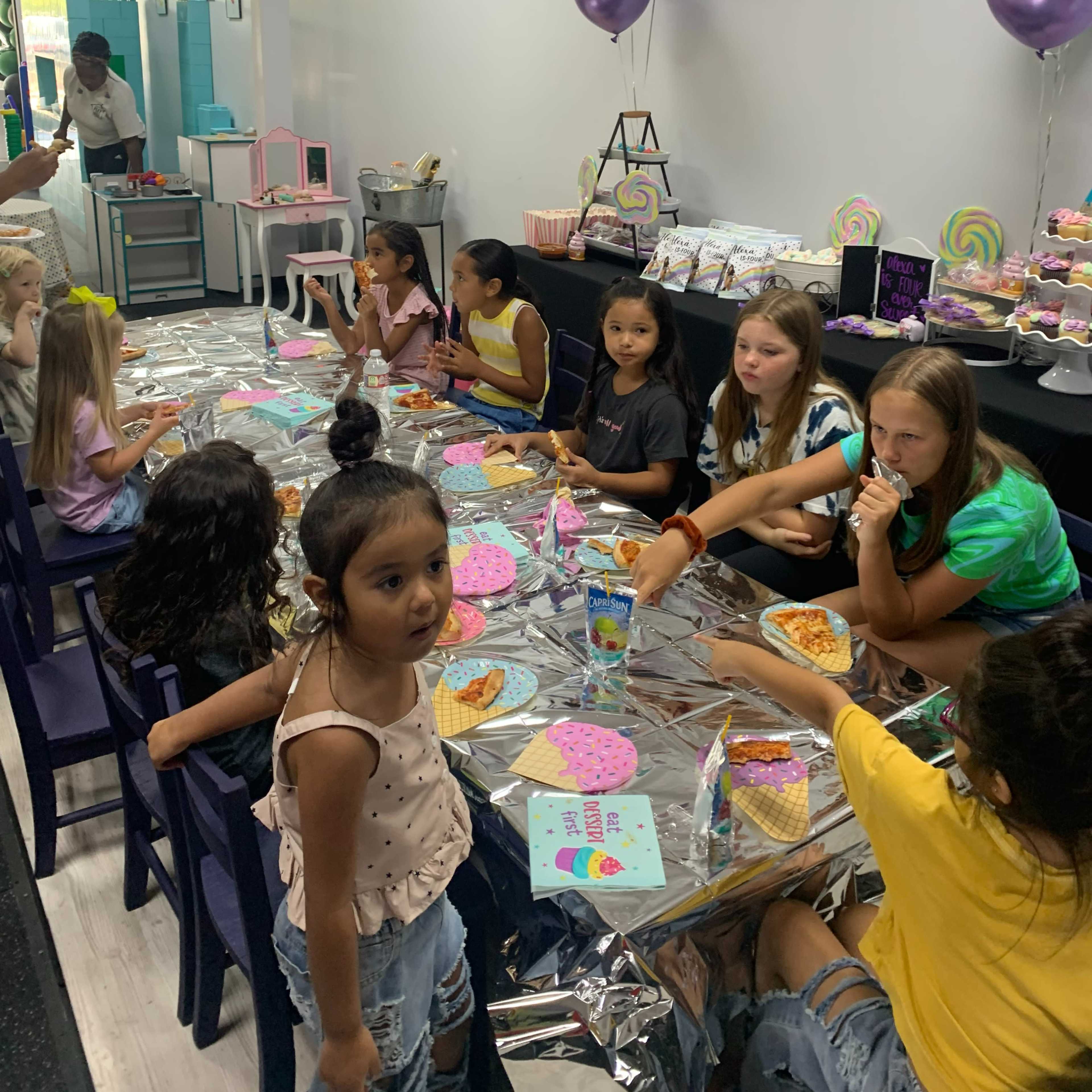 A group of children sits at a long table covered with colorful decorations, enjoying snacks and crafting activities at a festive indoor party.