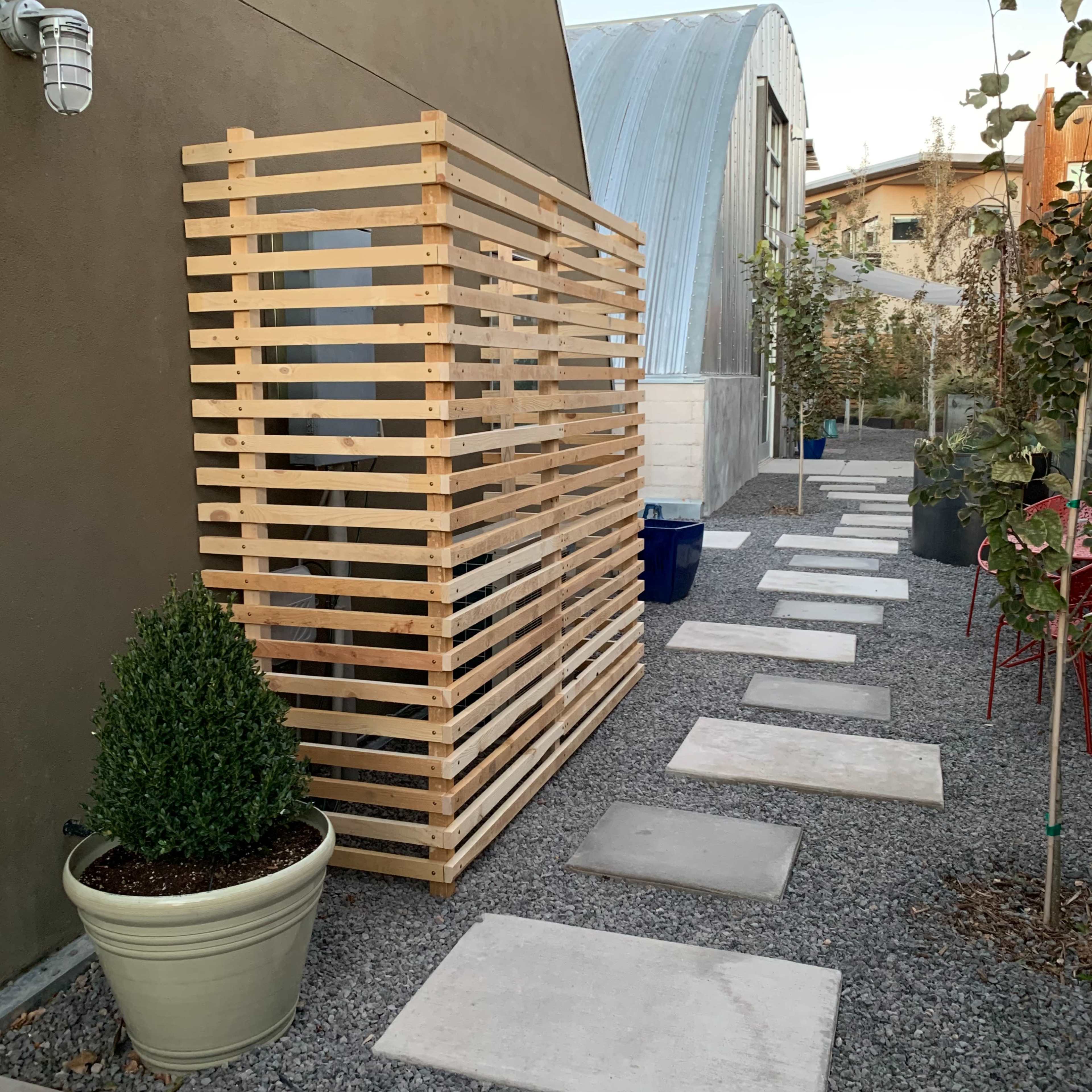 A wooden slatted structure stands beside a gravel pathway lined with concrete stepping stones and potted plants.