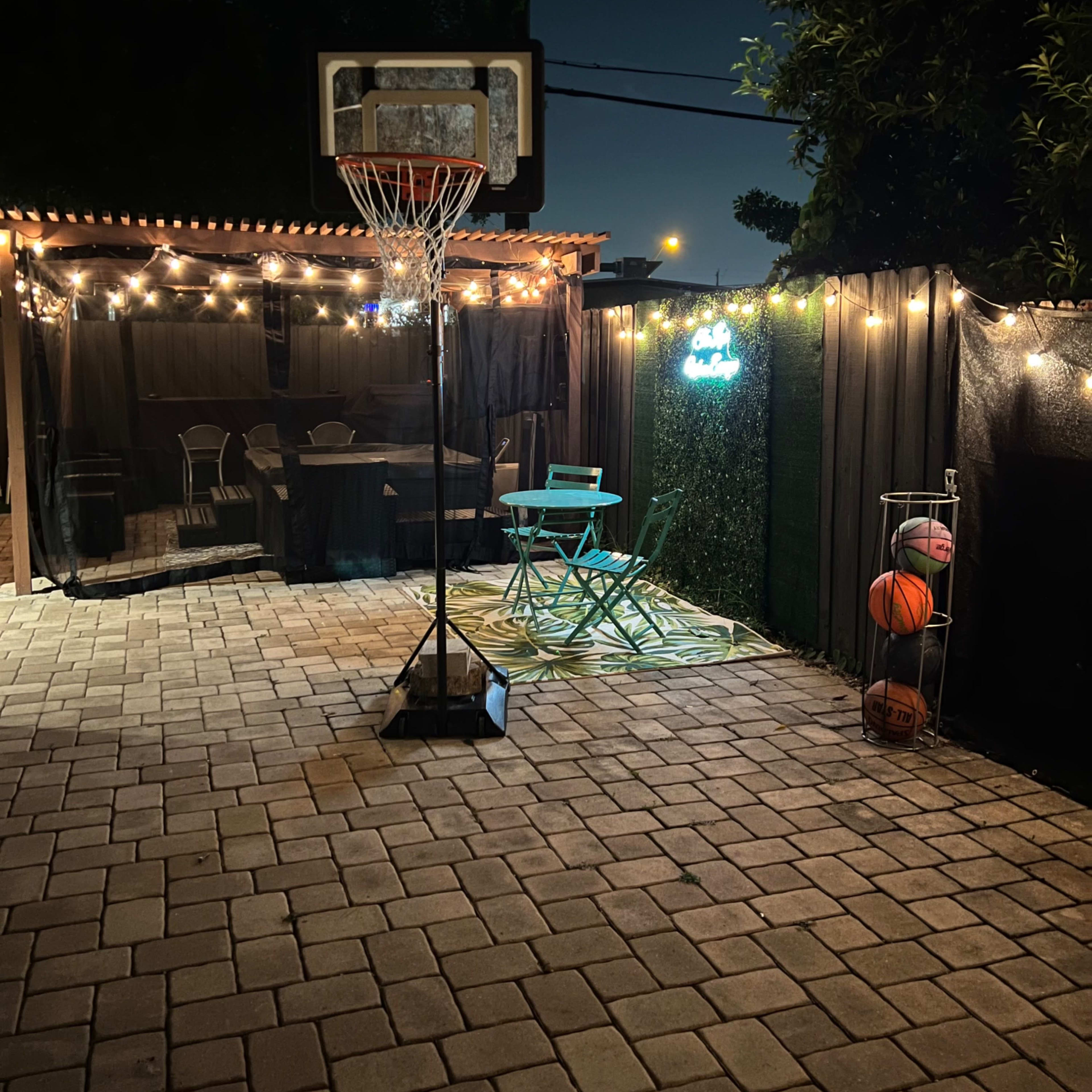 A backyard setup at night with a basketball hoop, string lights, a small table with chairs, and a basketball rack.