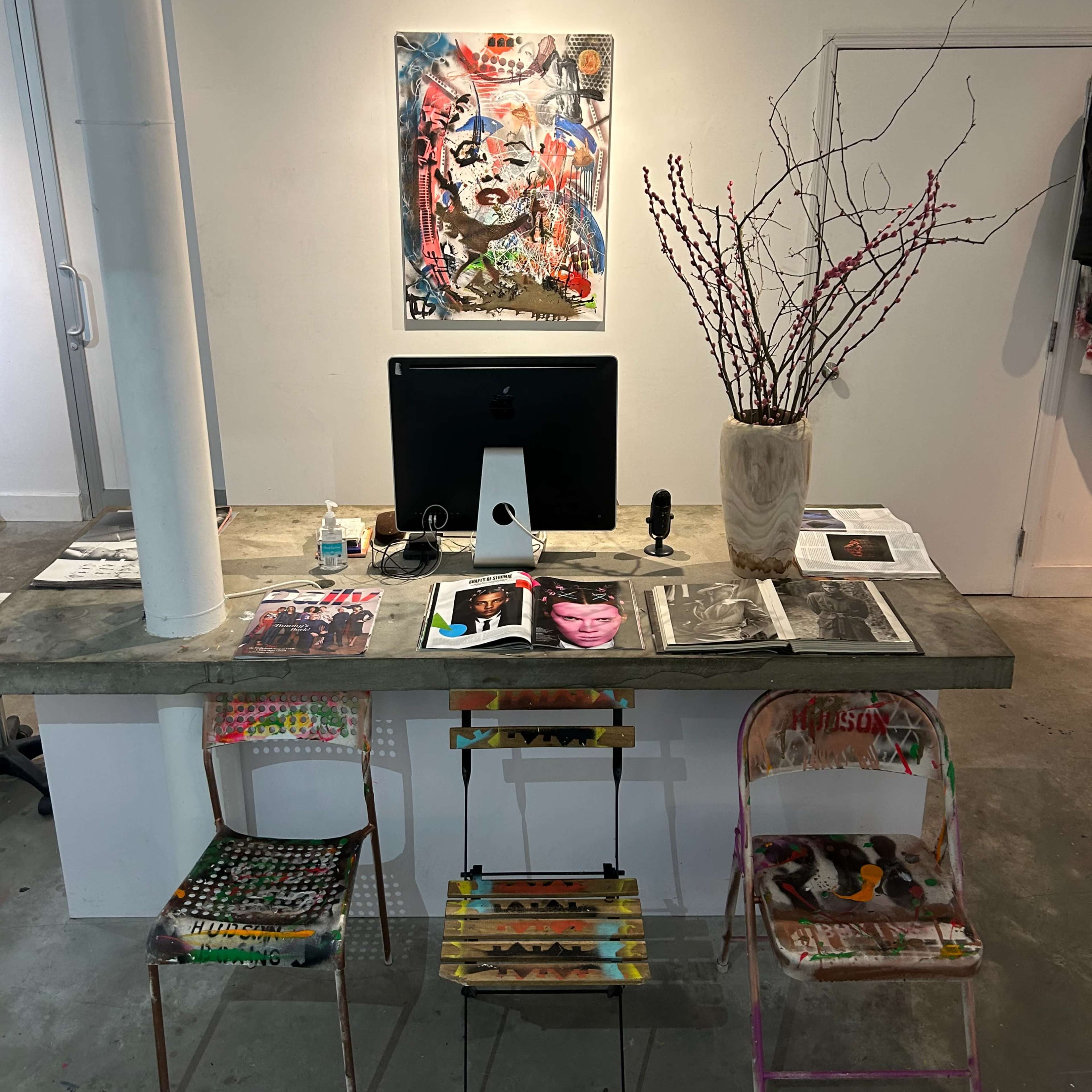 A minimalist workspace features a computer on a concrete desk, adorned with various printed materials and three colorful, painted chairs.