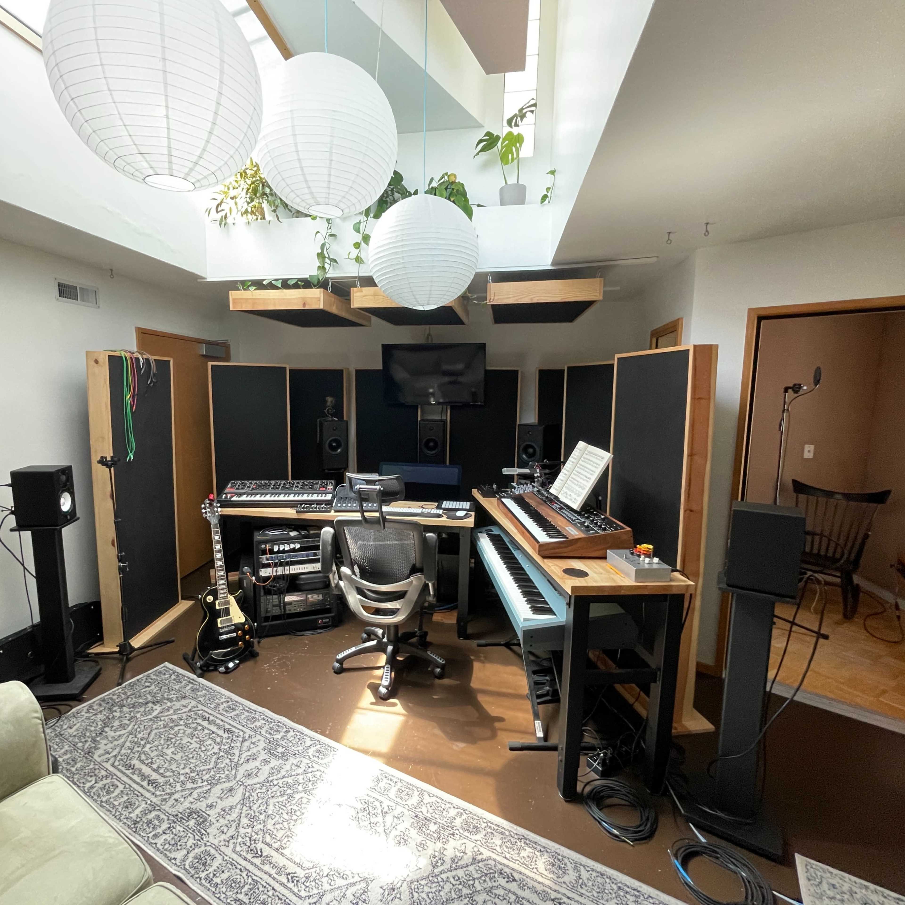 A modern recording studio with a central workspace featuring a mixing console, keyboard, and soundproofing panels, surrounded by seating and decorative pendant lights.