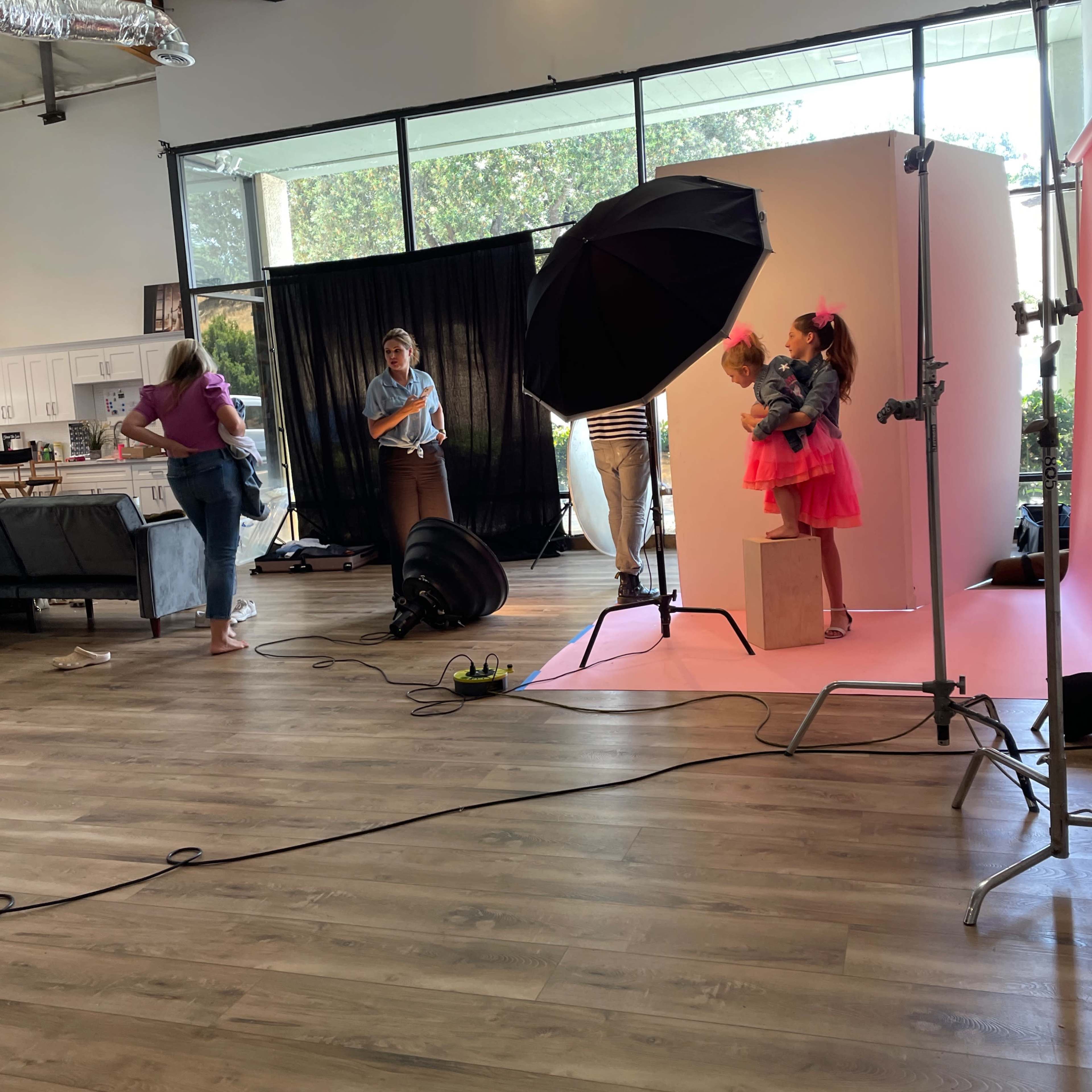A photoshoot is taking place in a spacious studio with a young girl in a pink outfit standing on a pedestal and several adults in the background.
