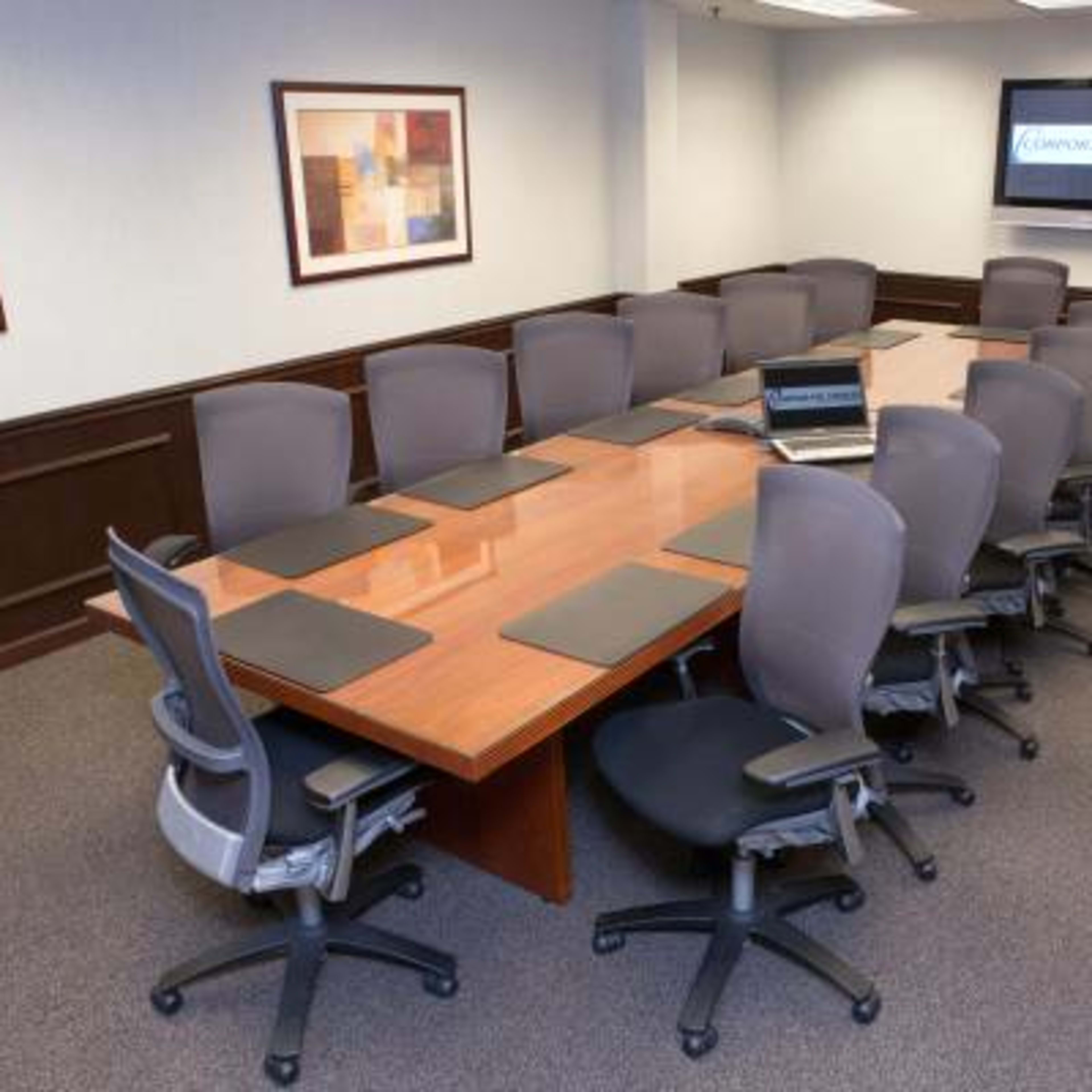 A large conference room features a long wooden table surrounded by ergonomic chairs, with an empty laptop placed on the table and a screen displaying a logo on the wall.