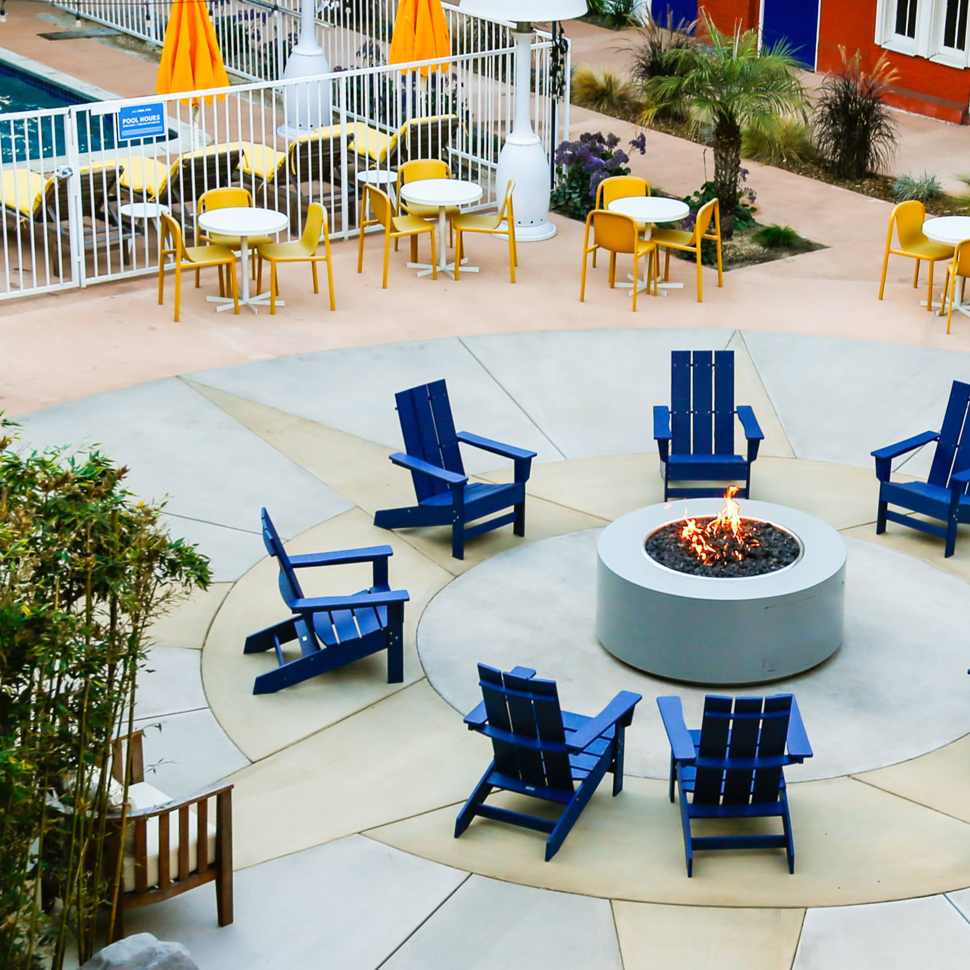 A circular arrangement of blue adirondack chairs surrounds a fire pit in a patio area next to a swimming pool.