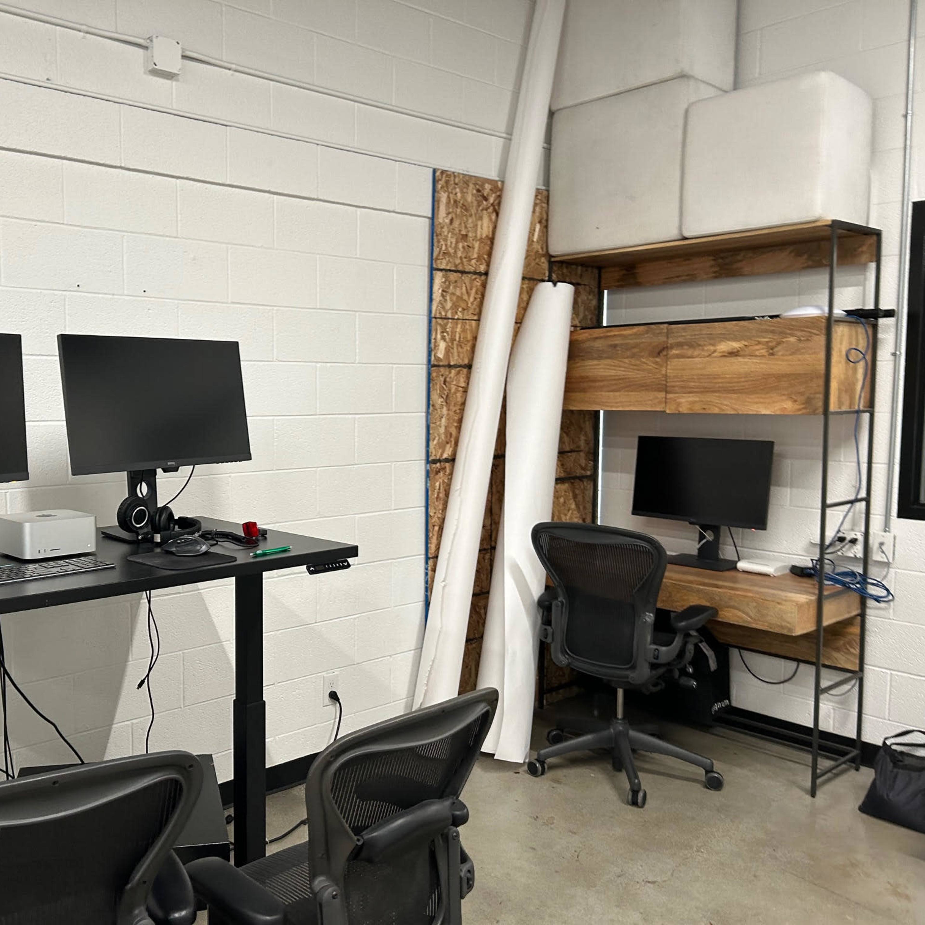A workspace with two monitors on a black standing desk, a desktop computer, and a smaller monitor on a wooden shelf, with two office chairs.