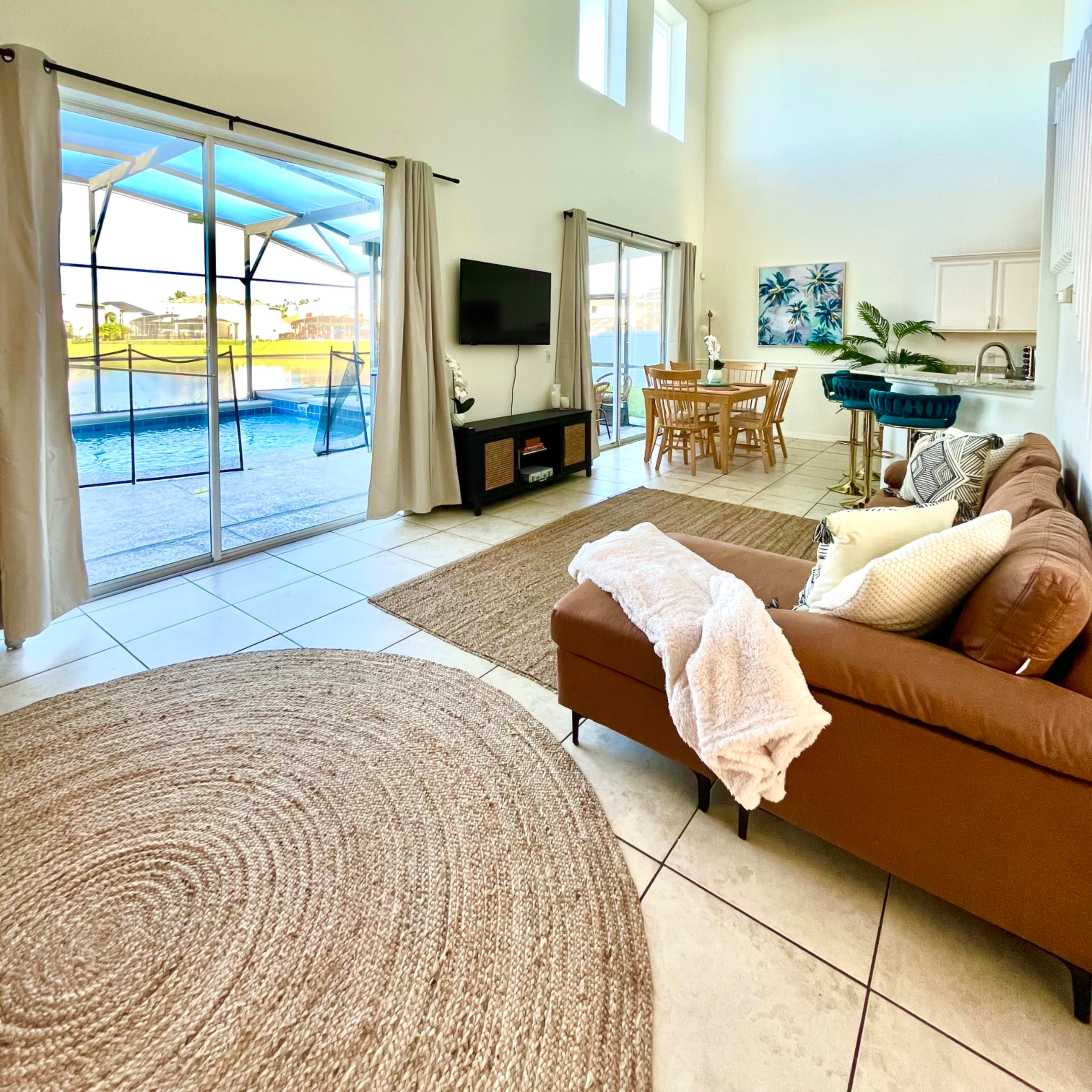 A spacious living room with a brown sofa, a round rug, a dining area, and large windows offering a view of an outdoor pool.