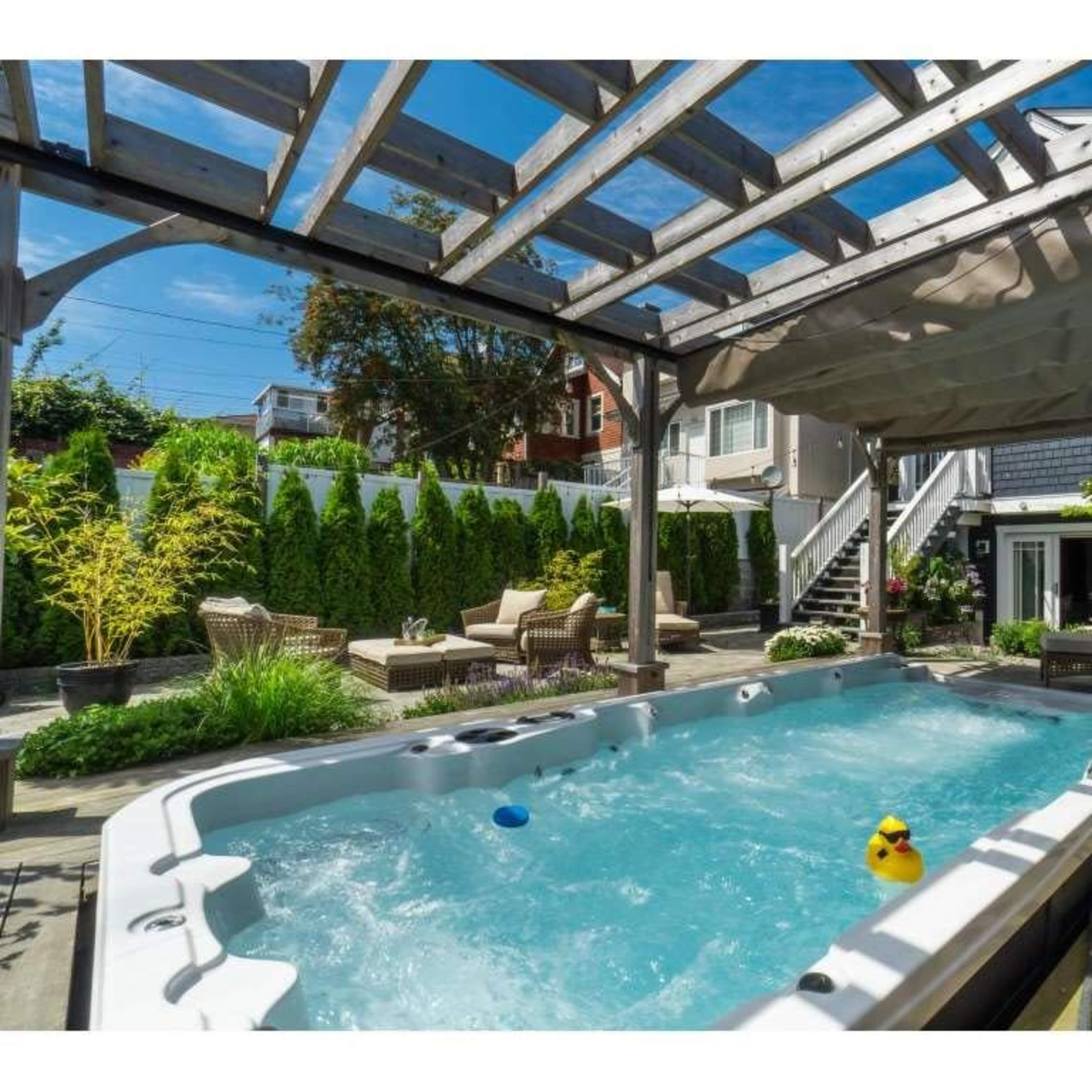 A spacious outdoor area featuring a hot tub surrounded by greenery, seating areas, and a wooden pergola.