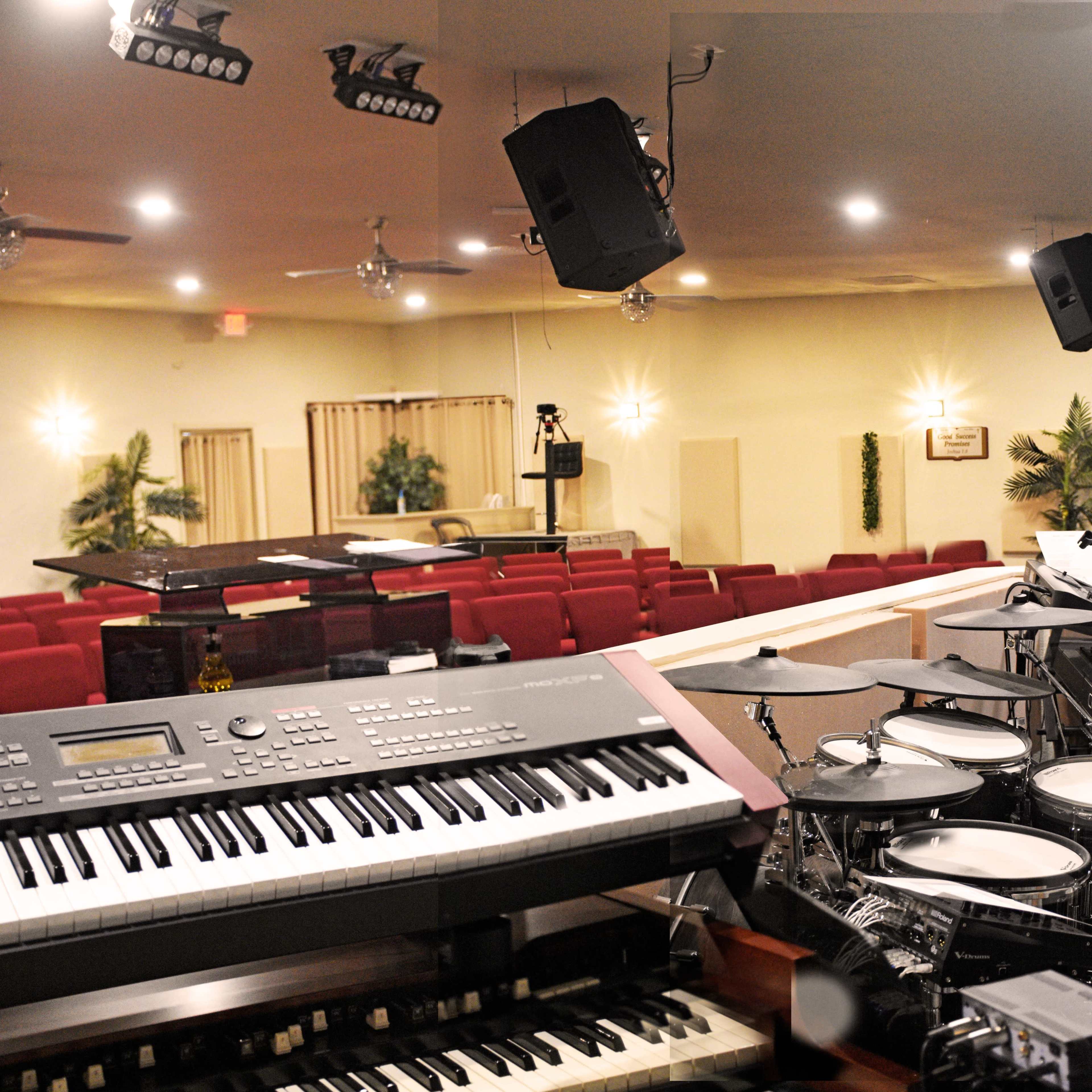 A music setup with electronic keyboards and a drum set positioned in front of an empty auditorium with red chairs.