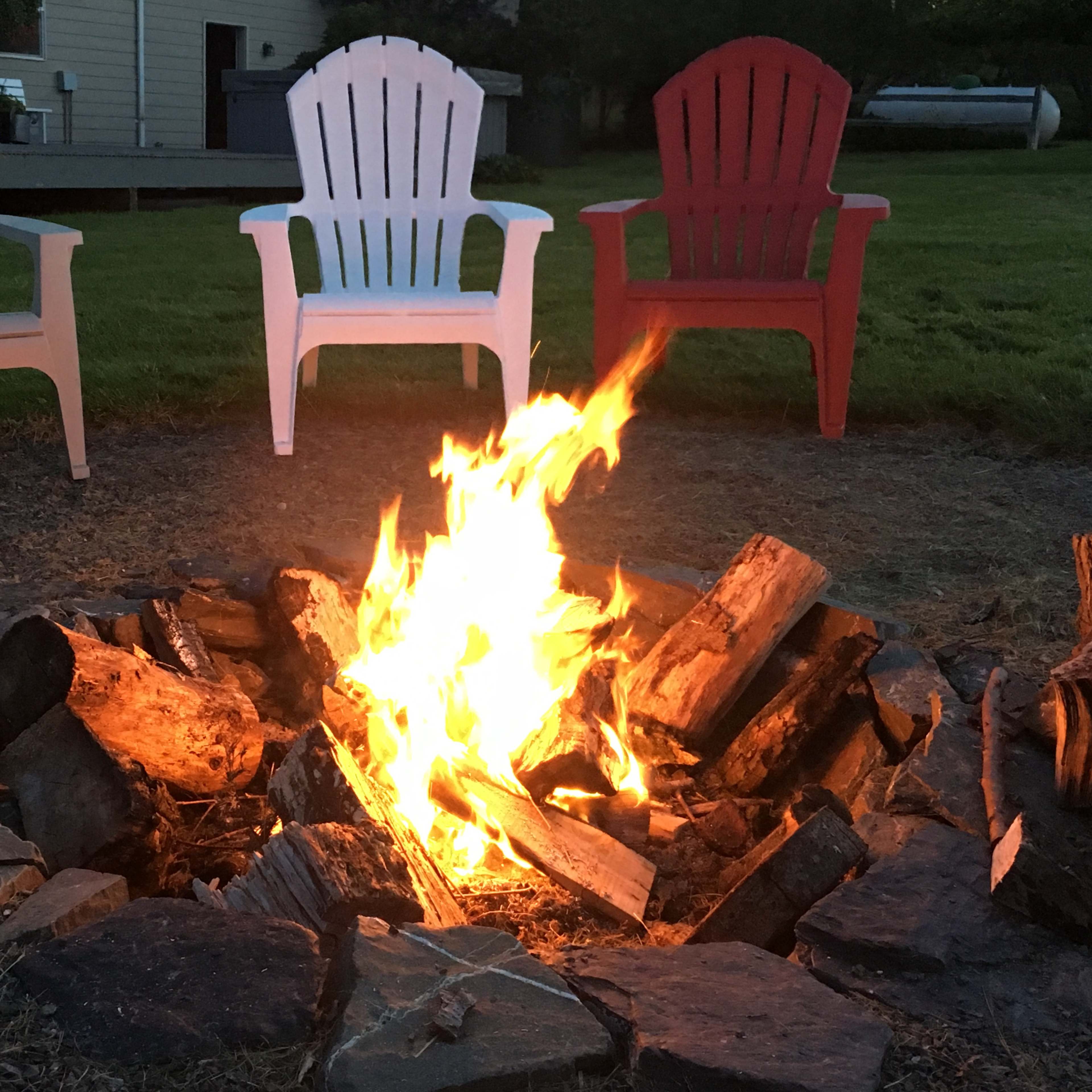 A fire burns in a stone circle surrounded by three Adirondack chairs in a backyard setting.
