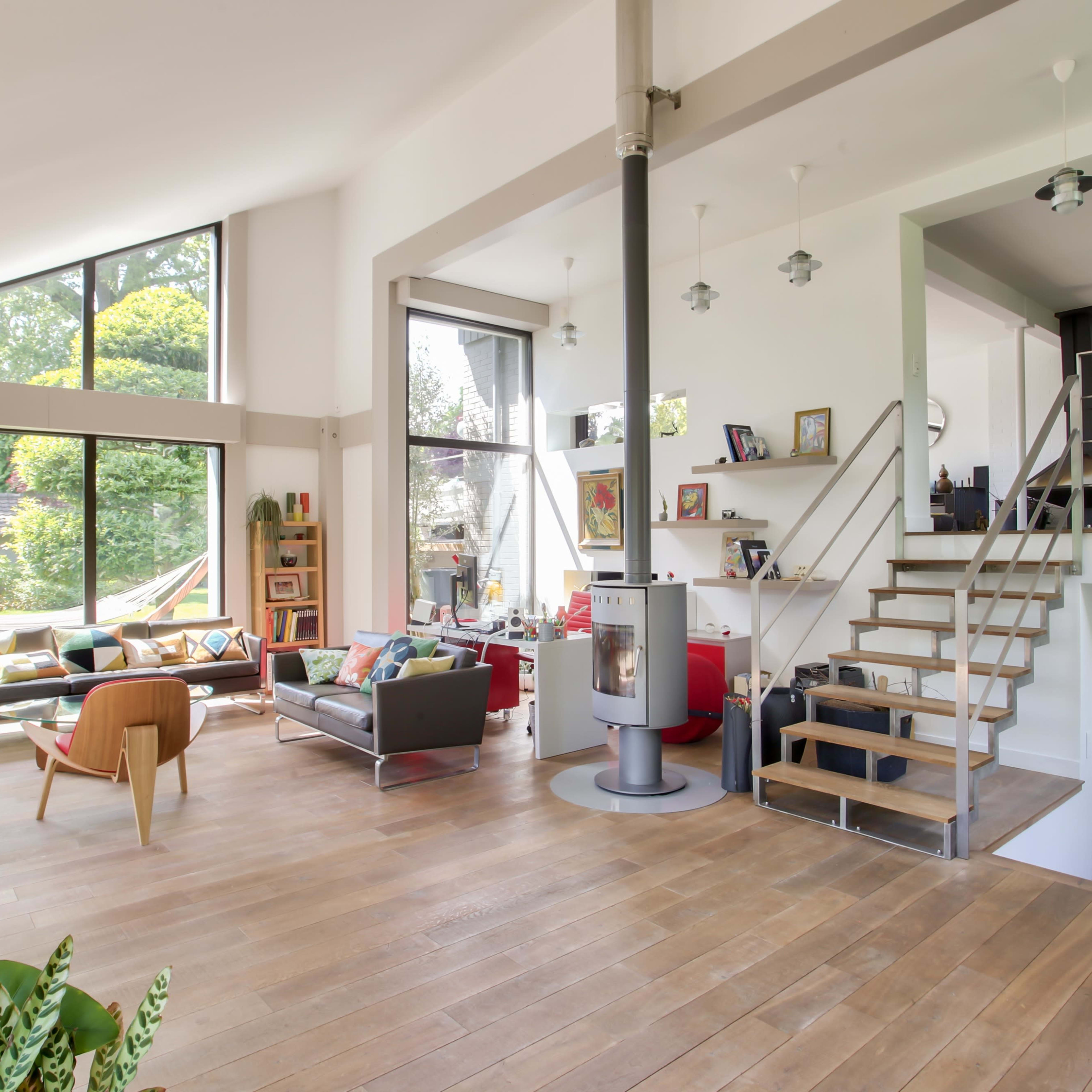The interior of a spacious, modern living room features large windows with a view of a garden, a staircase, and a mix of furniture including couches and chairs.