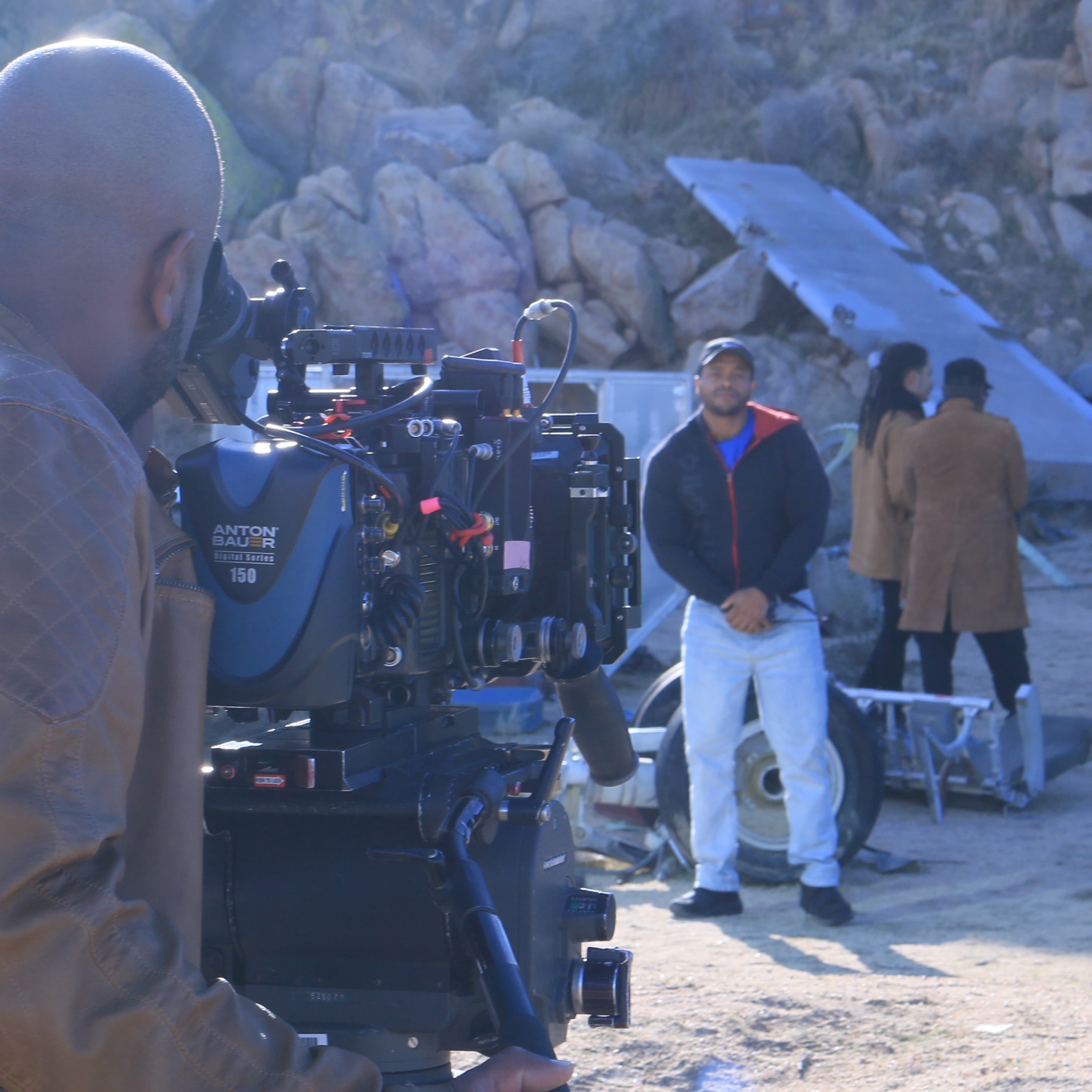A filmmaker operates a camera while a group of people stands in the background amidst rocky terrain and scattered equipment.