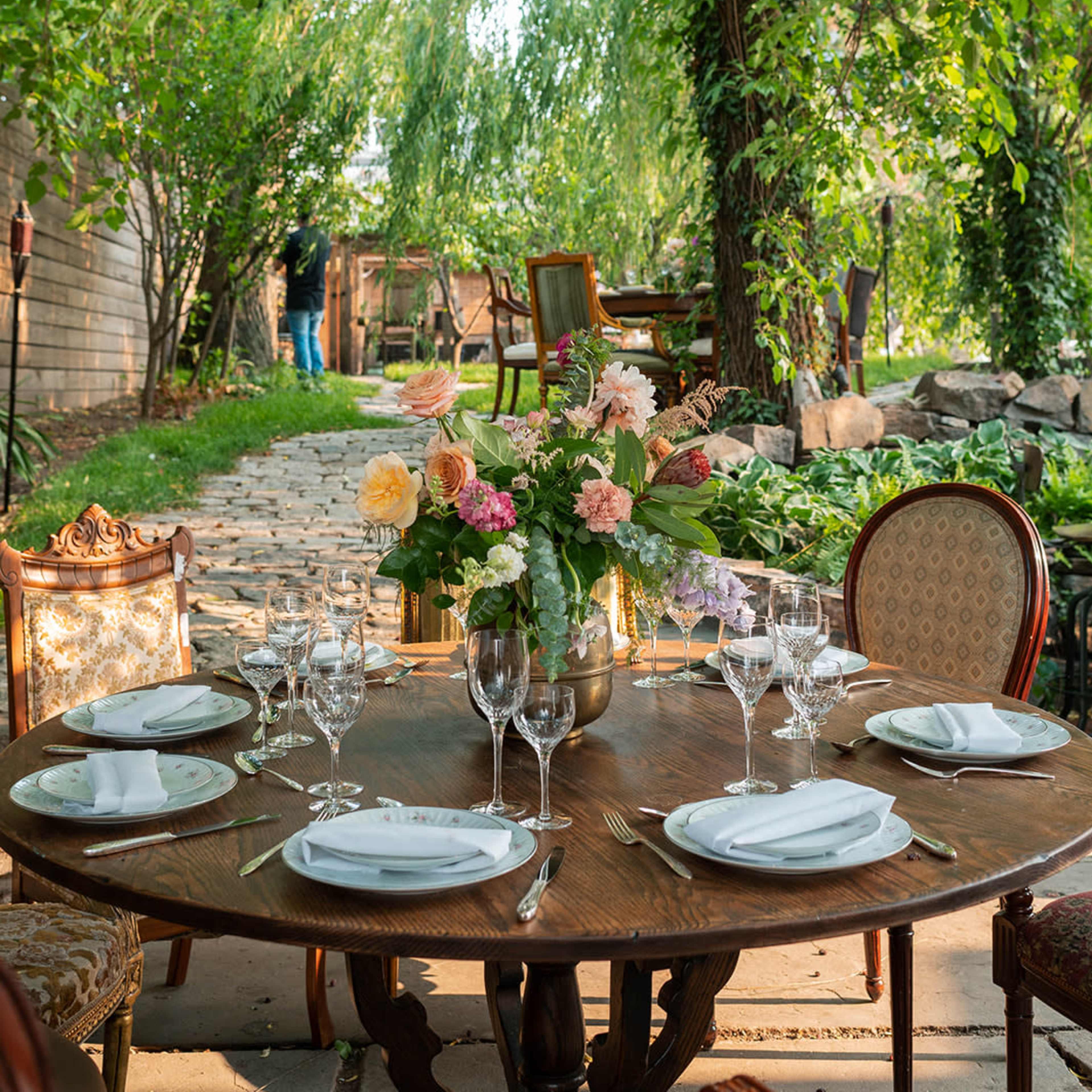 A beautifully set outdoor dining table with a chandelier overhead, surrounded by lush greenery and flowers.