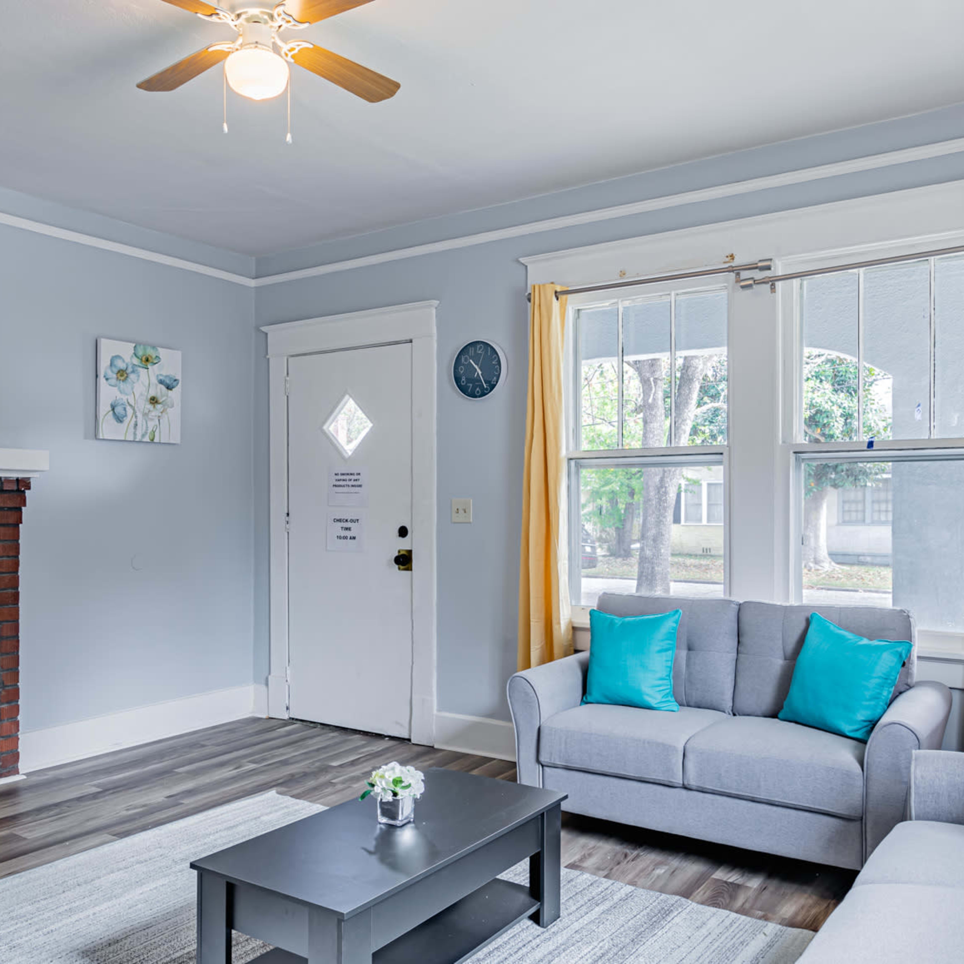 The living room features a gray sofa with teal cushions, a coffee table, a wall-mounted TV, and large windows with yellow curtains.