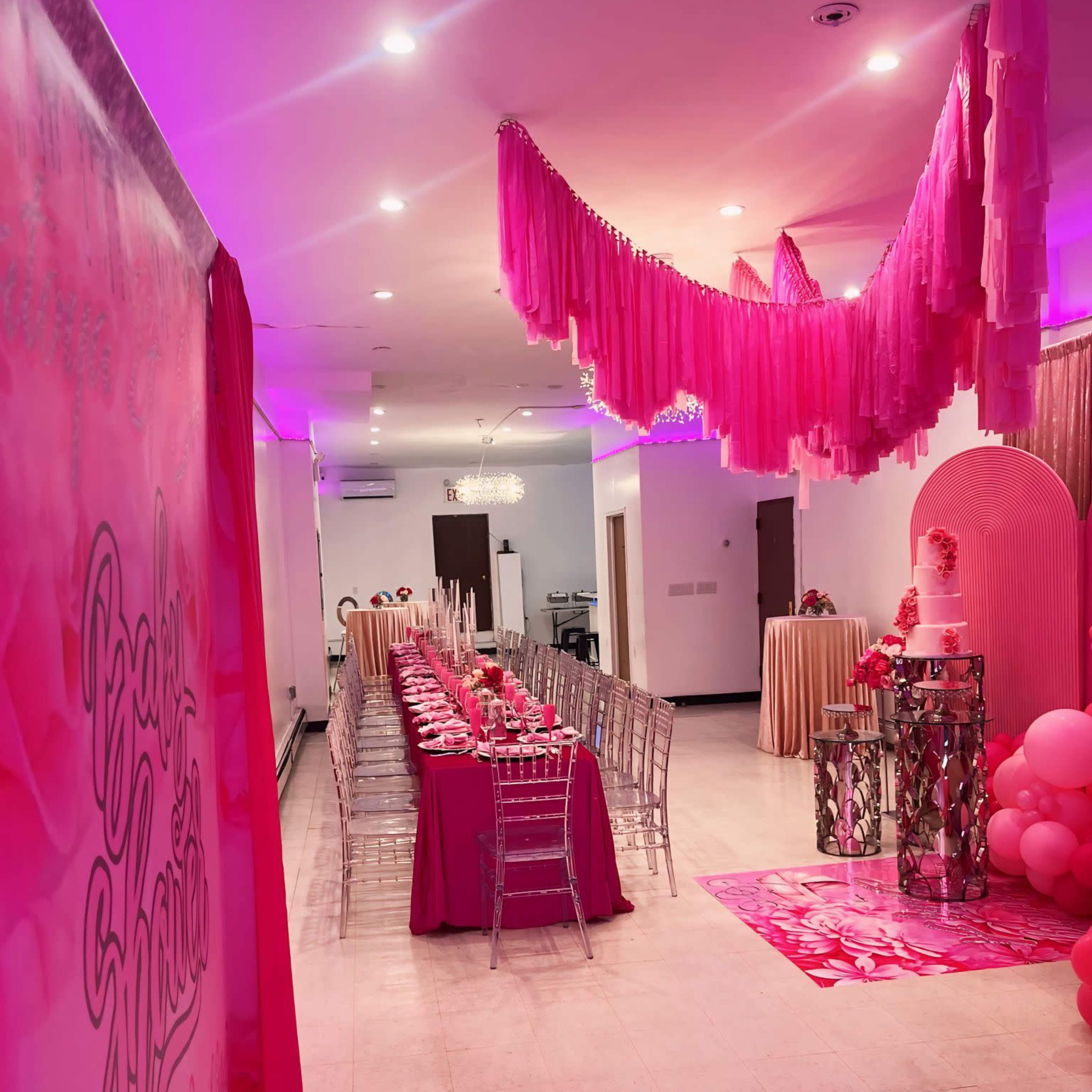 A brightly lit event space is decorated in shades of pink, featuring a long table set for a celebration, with streamers hanging from the ceiling and a cake displayed on a pedestal.