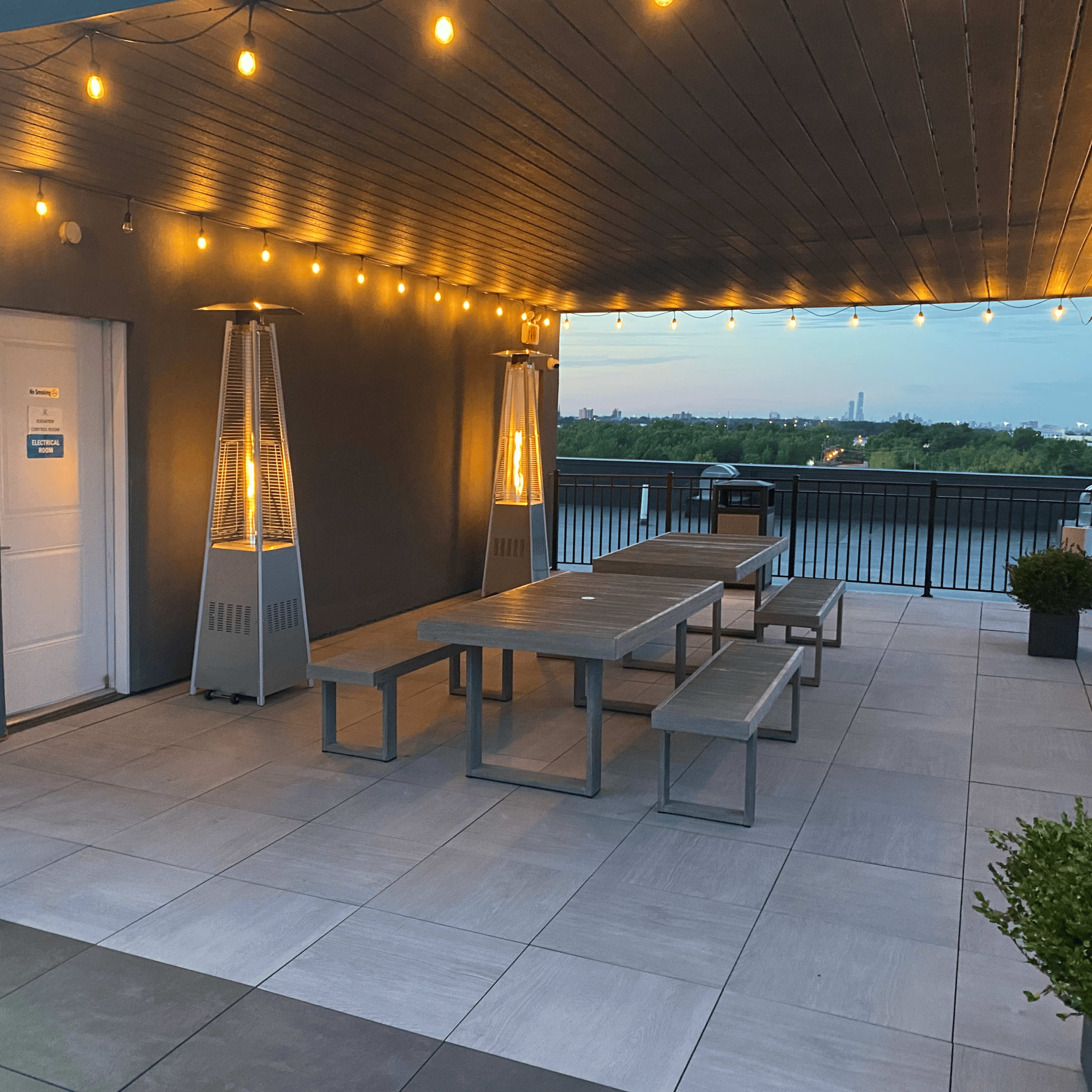 A rooftop terrace with string lights, two outdoor heaters, and a long table with benches overlooking a body of water.