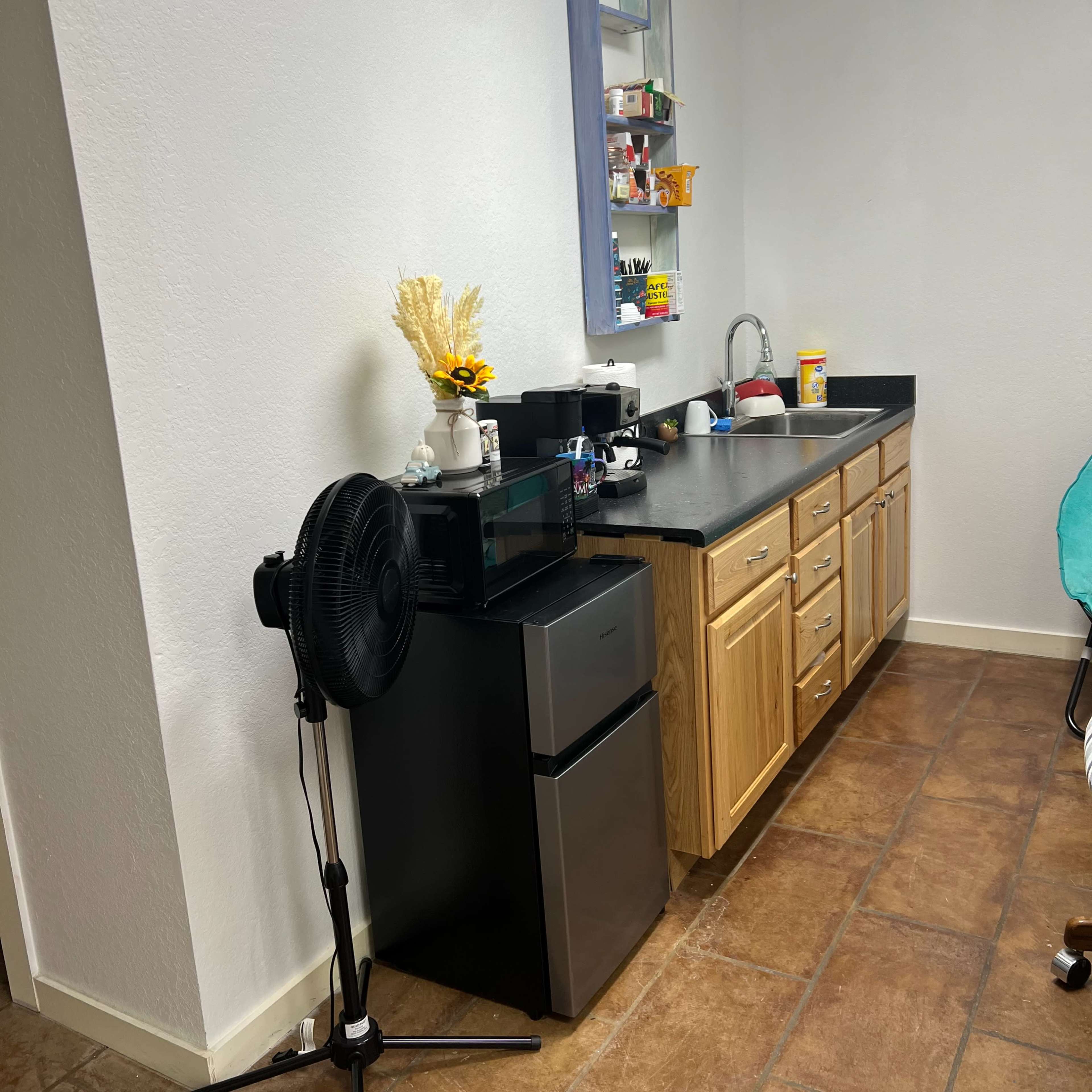 A small kitchen area featuring a black microwave and a compact refrigerator next to wooden cabinets with a sink and various items on the countertop.