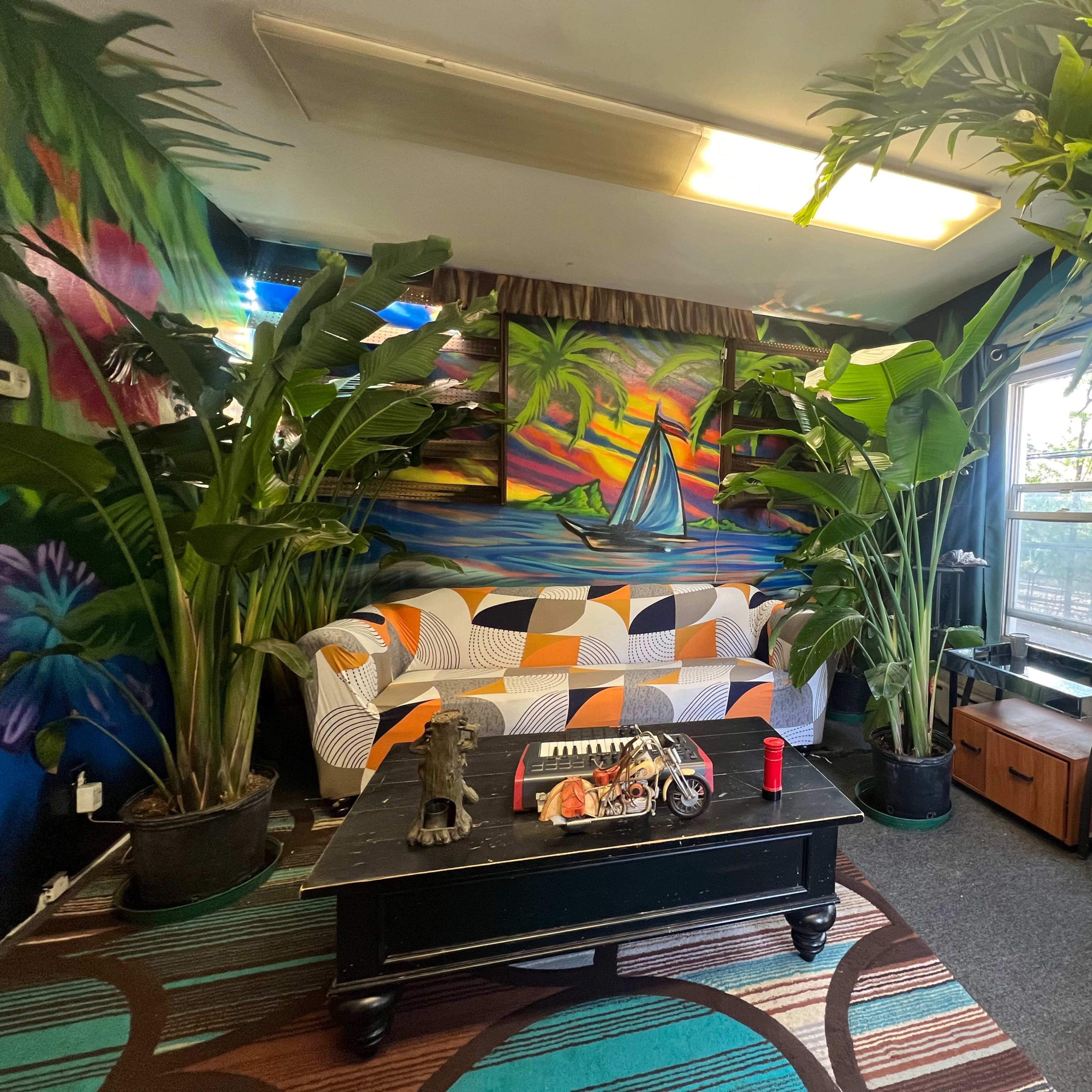 A colorful room with a patterned sofa, a black coffee table adorned with items, and large potted plants beside a mural featuring a sailboat on water.