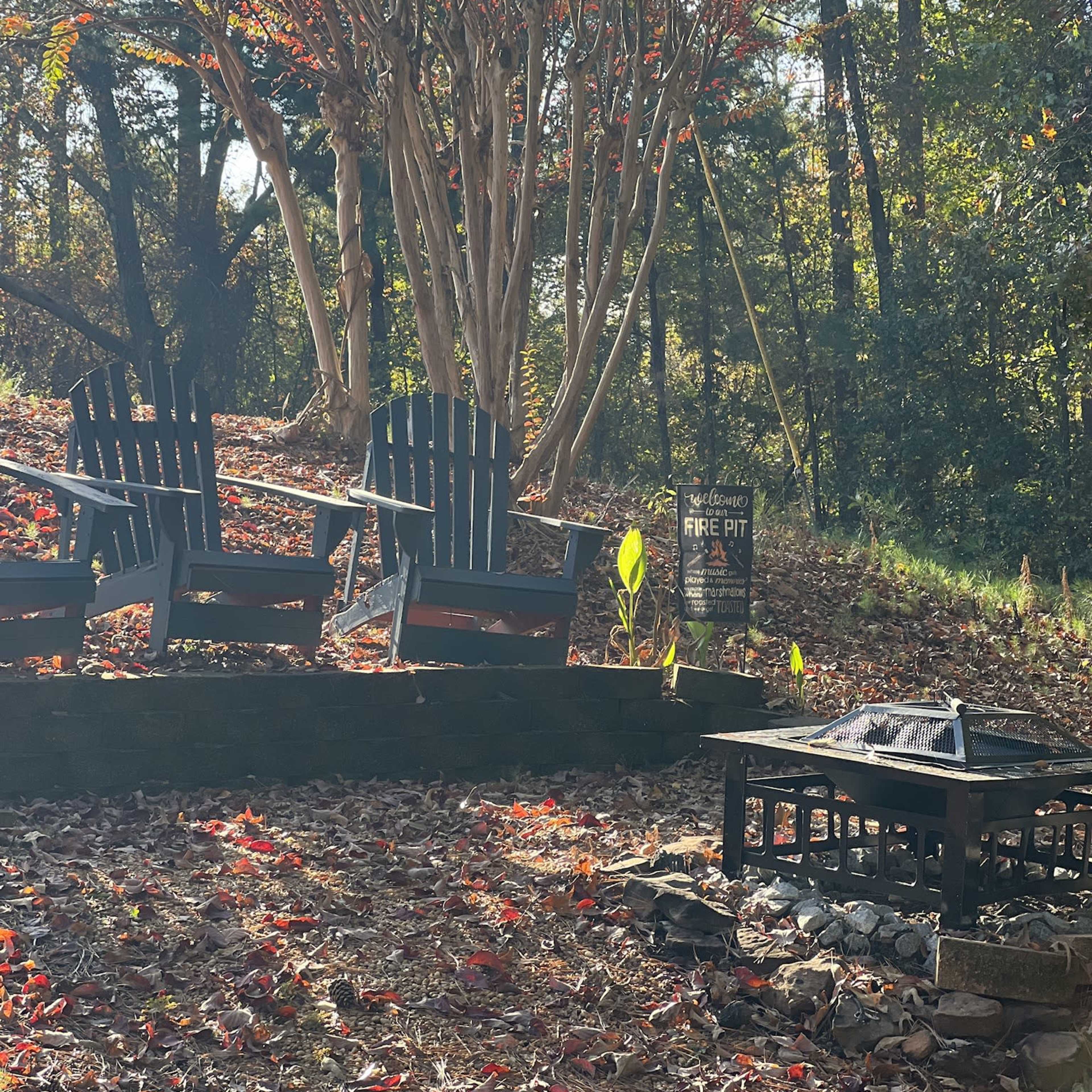 A cozy outdoor space features three black adirondack chairs facing a stone fire pit, surrounded by autumn leaves and a small tree with red foliage.