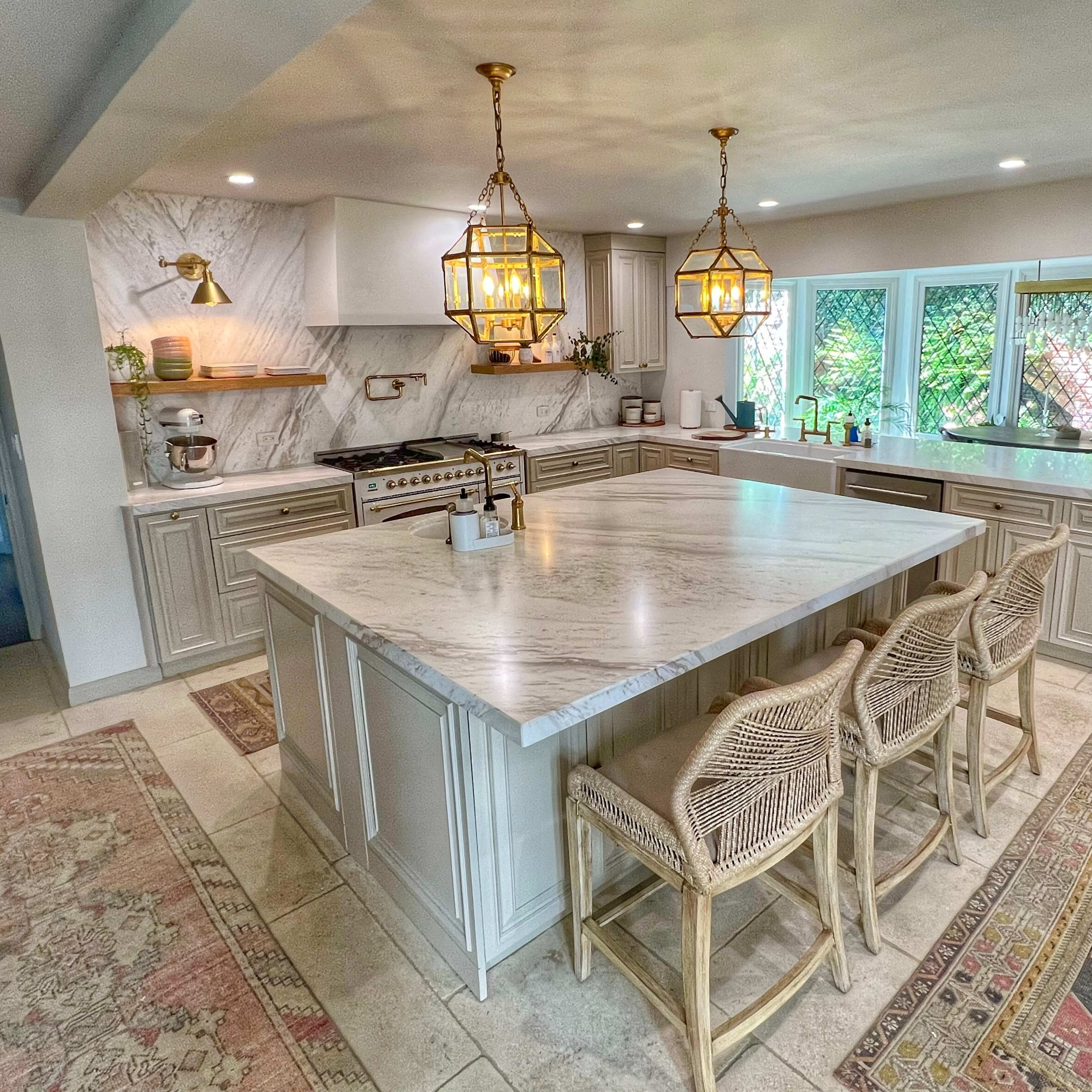 The kitchen features a large marble island with four rattan bar stools, stylish pendant lights, and modern appliances among light cabinetry and natural light coming from surrounding windows.