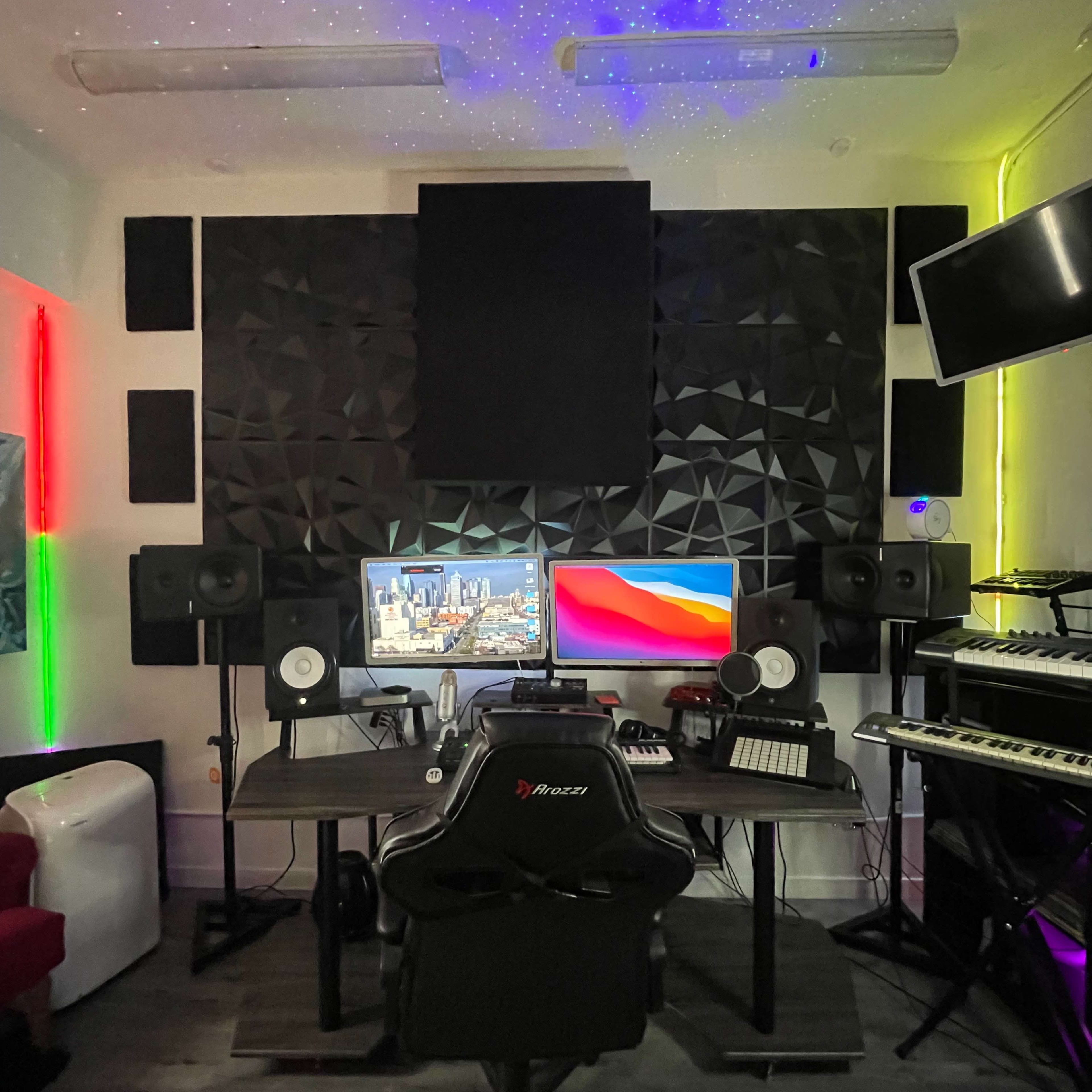 A music production studio featuring two computer monitors, surround speakers, a keyboard, and colorful LED lighting on the walls.