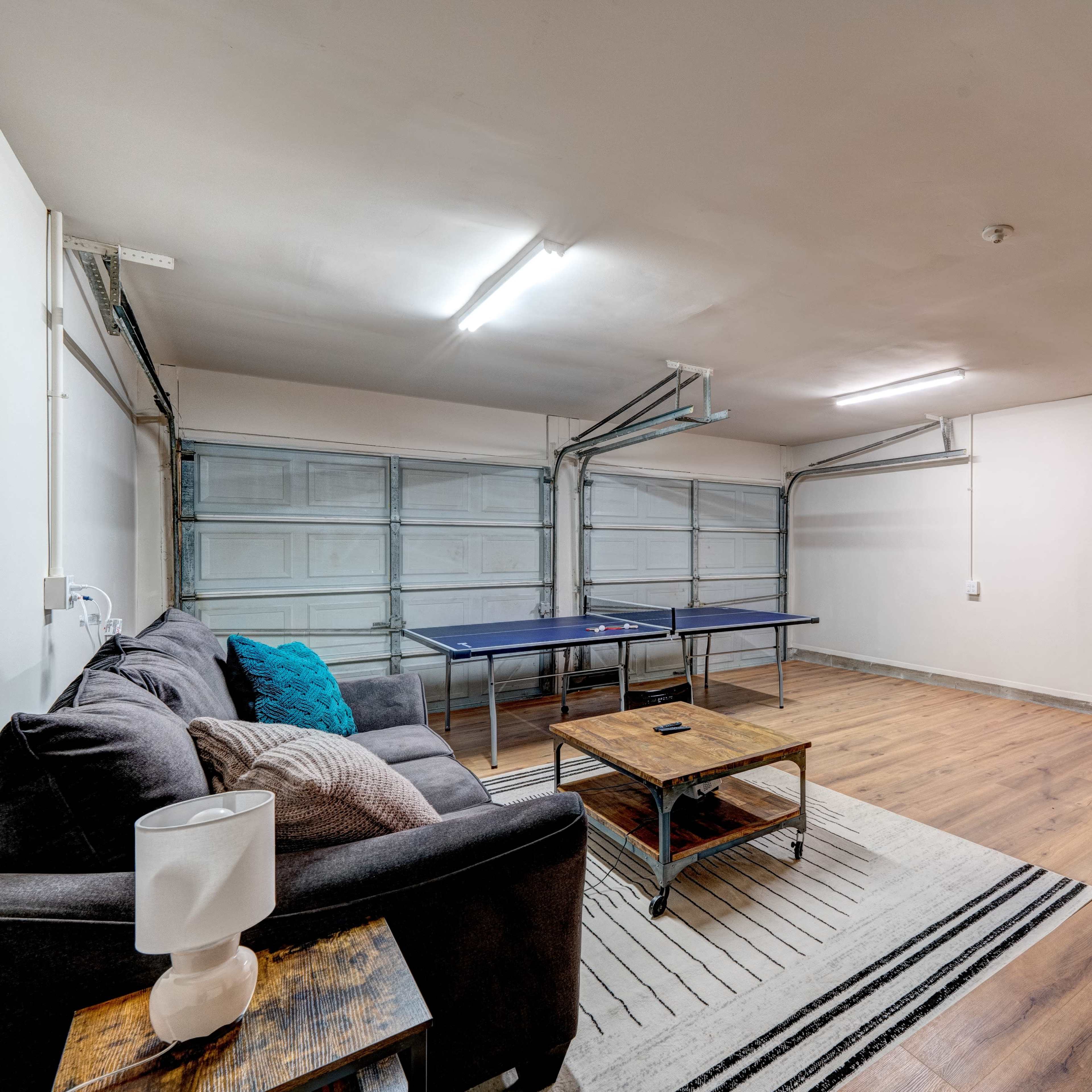 A clean garage features a couch, coffee table, and a ping pong table, with overhead lights illuminating the space.